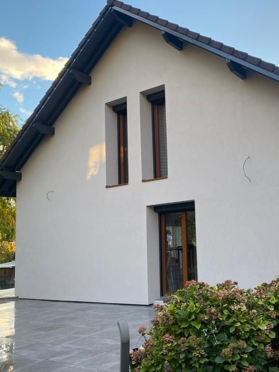 Maison blanche au toit foncé et aux fenêtres à encadrement marron, sur fond de ciel nuageux.
