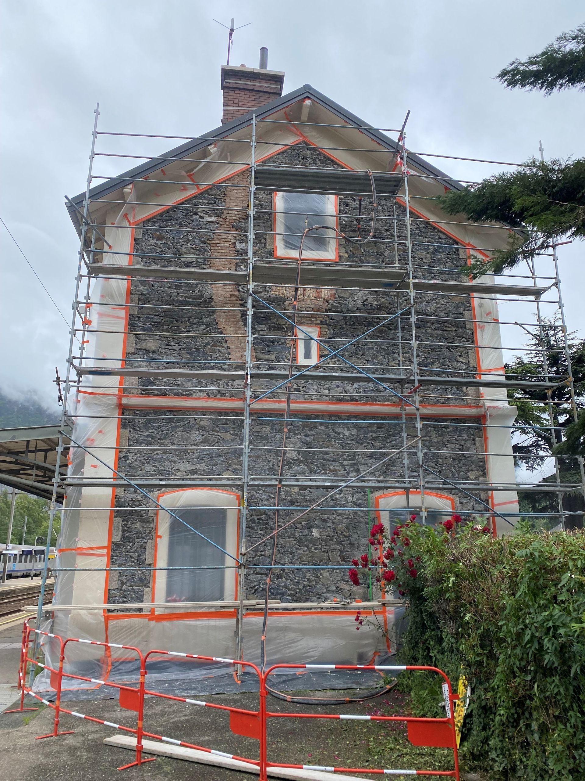 Façade d'une maison en rénovation avec échafaudages ; murs en pierre grise, boiseries orange.