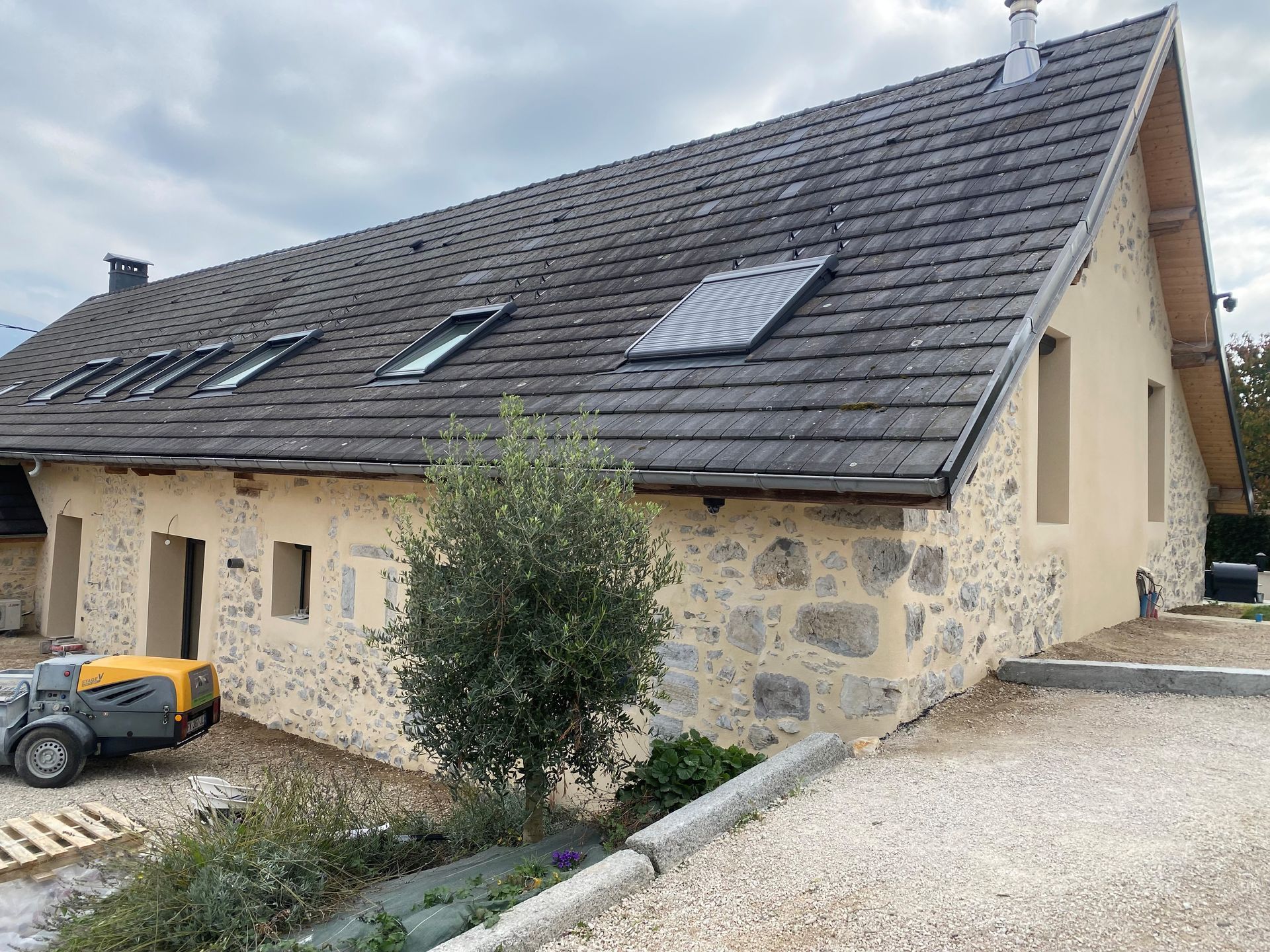 Bâtiment à façade en pierre, toit gris et puits de lumière. Olivier au premier plan.