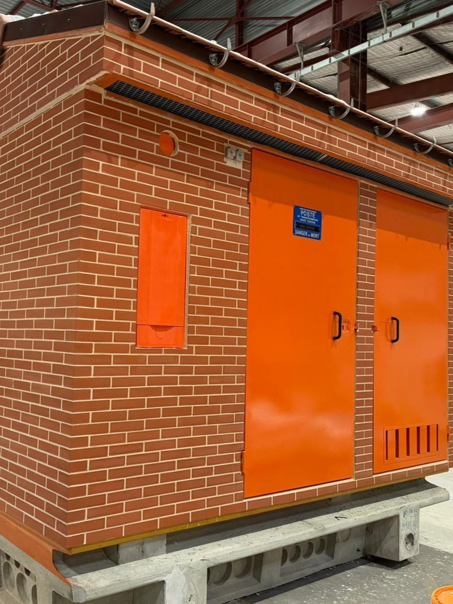 Bâtiment en briques avec portes et fenêtres orange. Il repose sur une base en béton, avec un toit sombre et une plaque sur la porte.