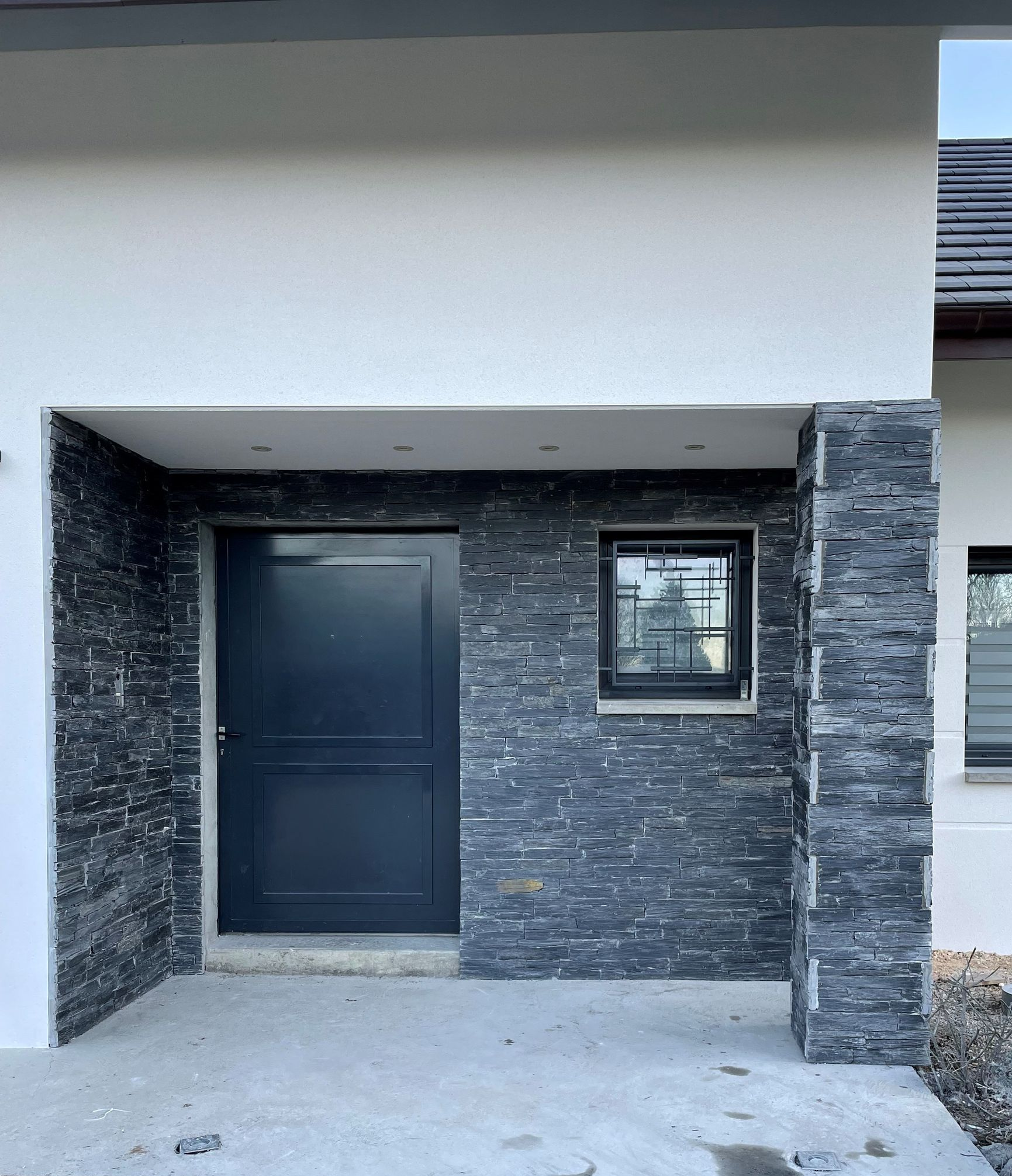 Entrée d'un bâtiment avec une porte noire et une façade en pierre sous un auvent blanc.