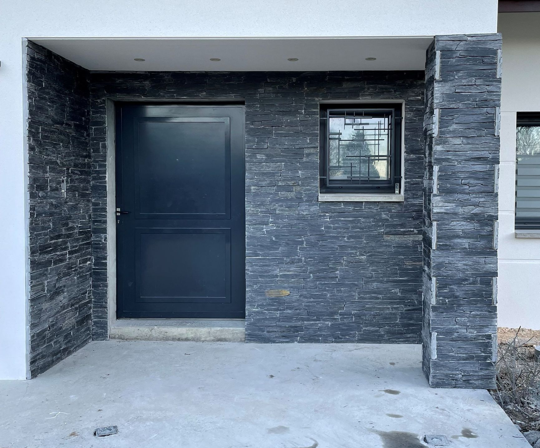 Porte d'entrée gris foncé et mur d'accent avec petite fenêtre et entrée en béton.