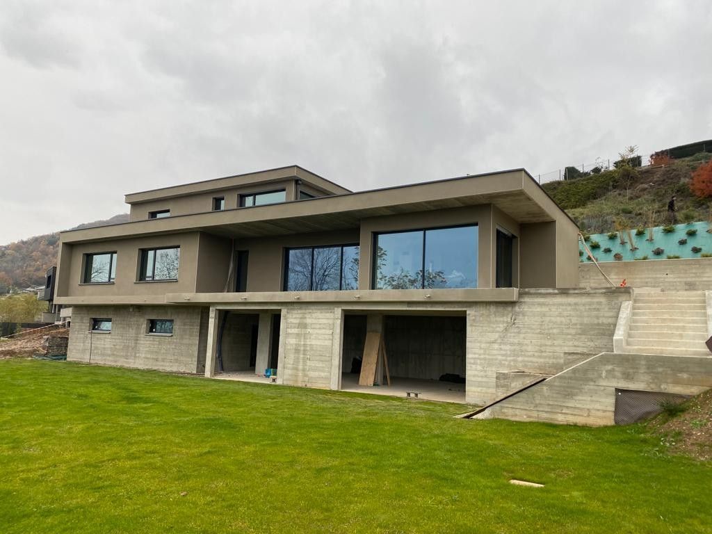 Maison moderne de deux étages en construction, avec de grandes fenêtres et des fondations en béton, sur un coteau arboré.
