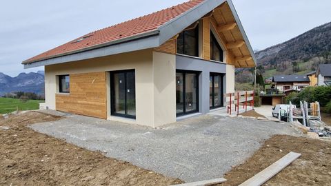 Maison neuve en construction, aux murs beiges et gris, avec des éléments en bois et un toit de tuiles rouges, le tout sur fond de montagnes.