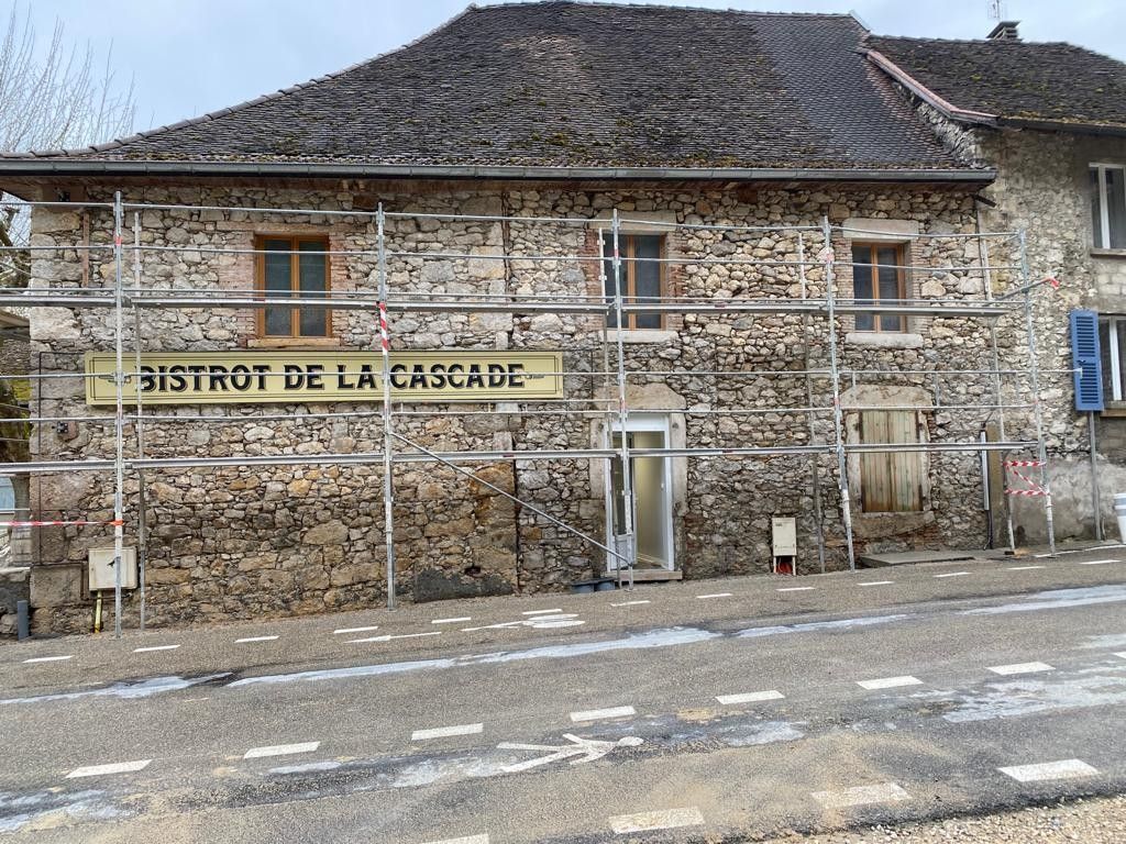 Bâtiment en pierre à trois fenêtres avec une enseigne « Bistrot de la Cascade », échafaudages et une route devant.