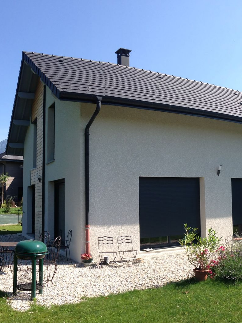 Maison à deux étages avec toit et garage gris foncé, murs beige clair et boiseries noires. Pelouse verte et gravier.