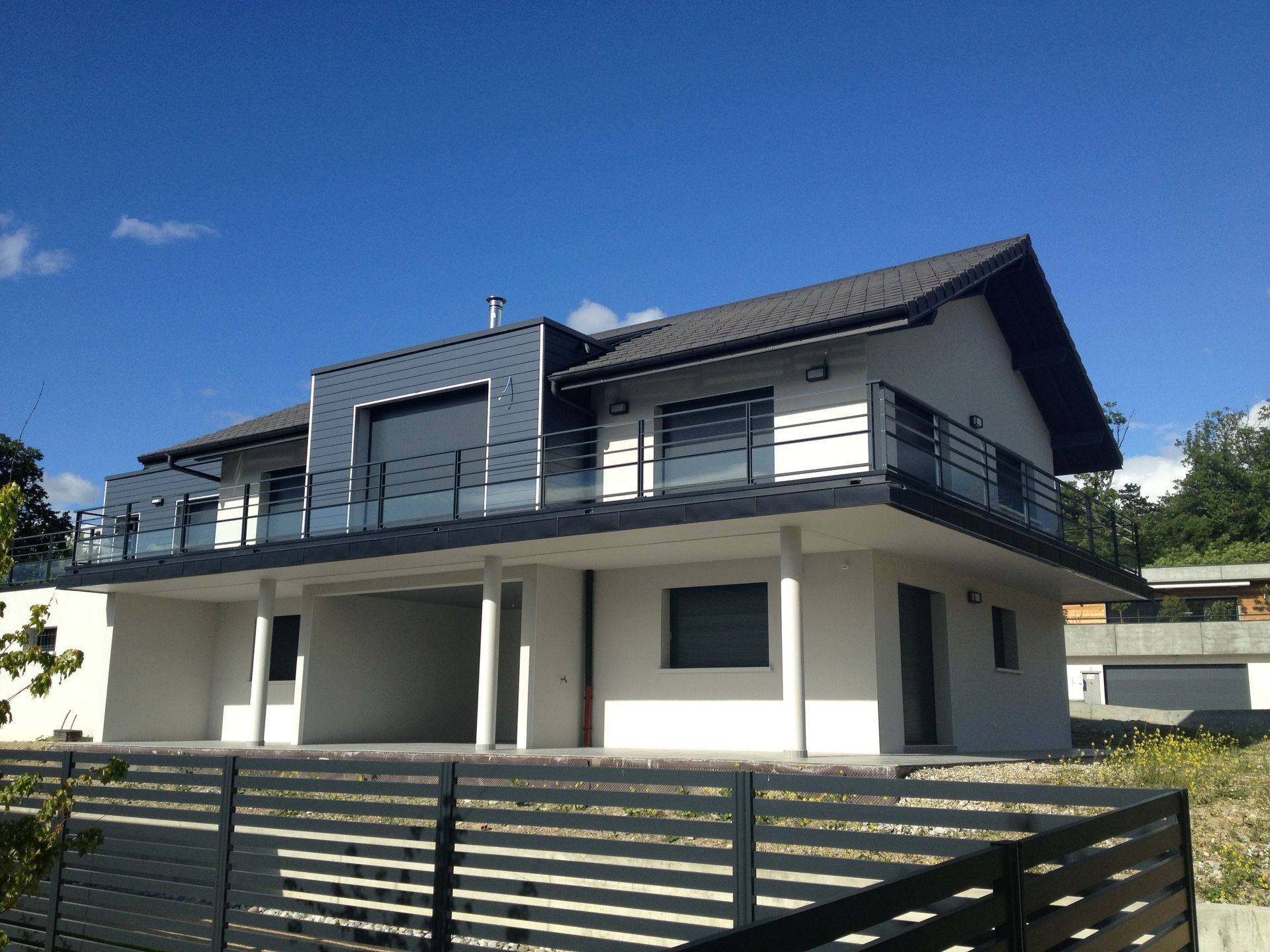 Maison moderne blanche et gris foncé à deux étages avec toit pentu et balcon sur fond de ciel bleu.