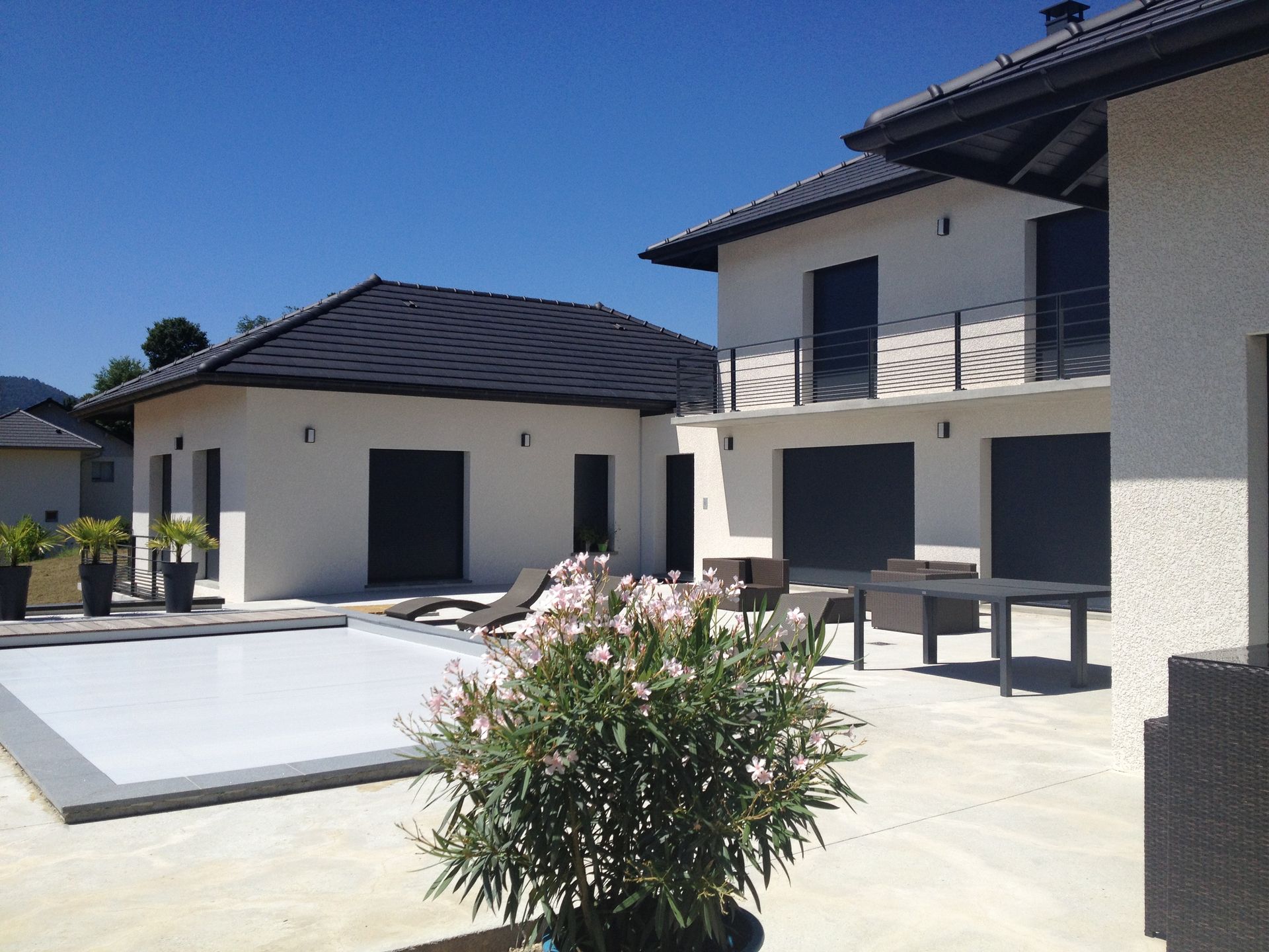 Maison blanche moderne avec volets et toit foncés, piscine et mobilier de terrasse sous un ciel bleu.