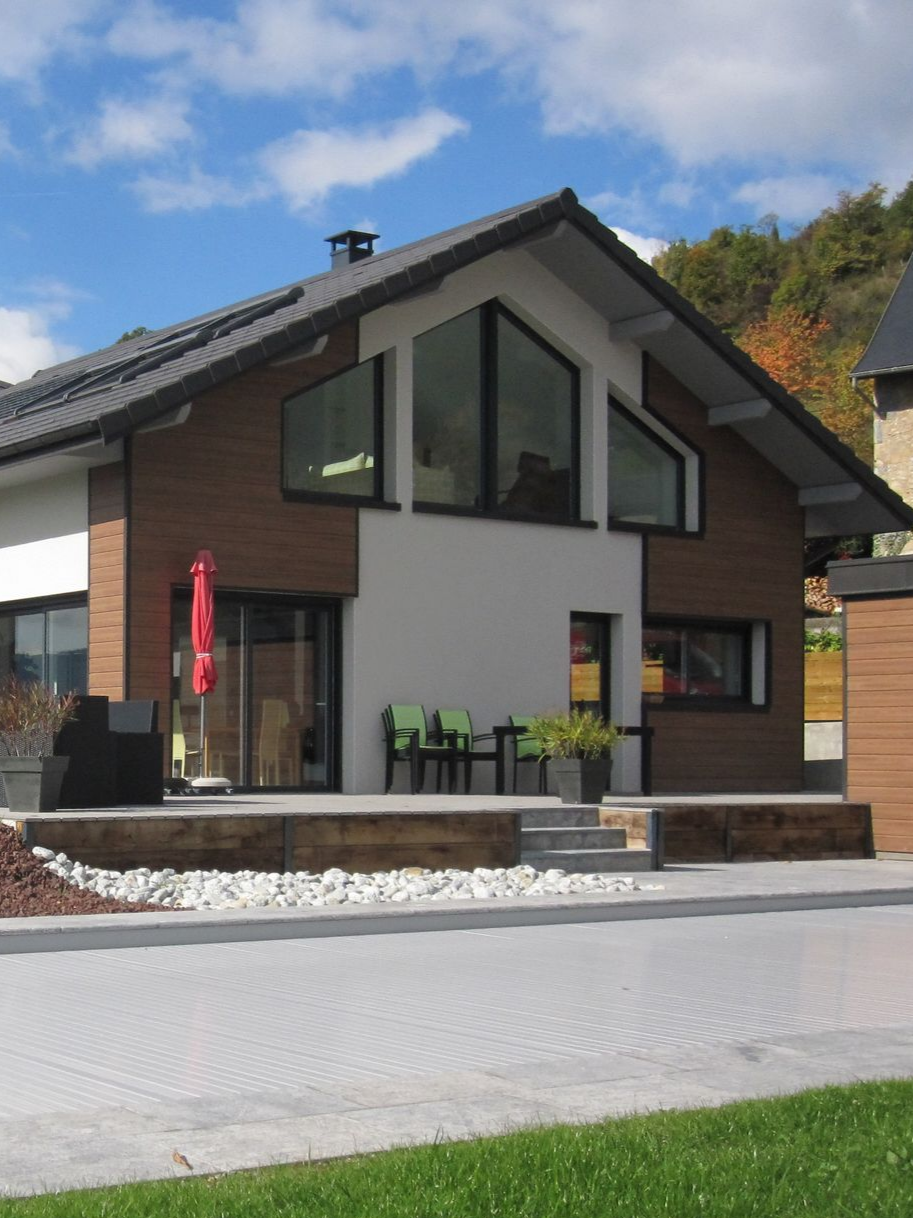 Maison moderne avec façade marron et blanche, grandes fenêtres et terrasse pavée.