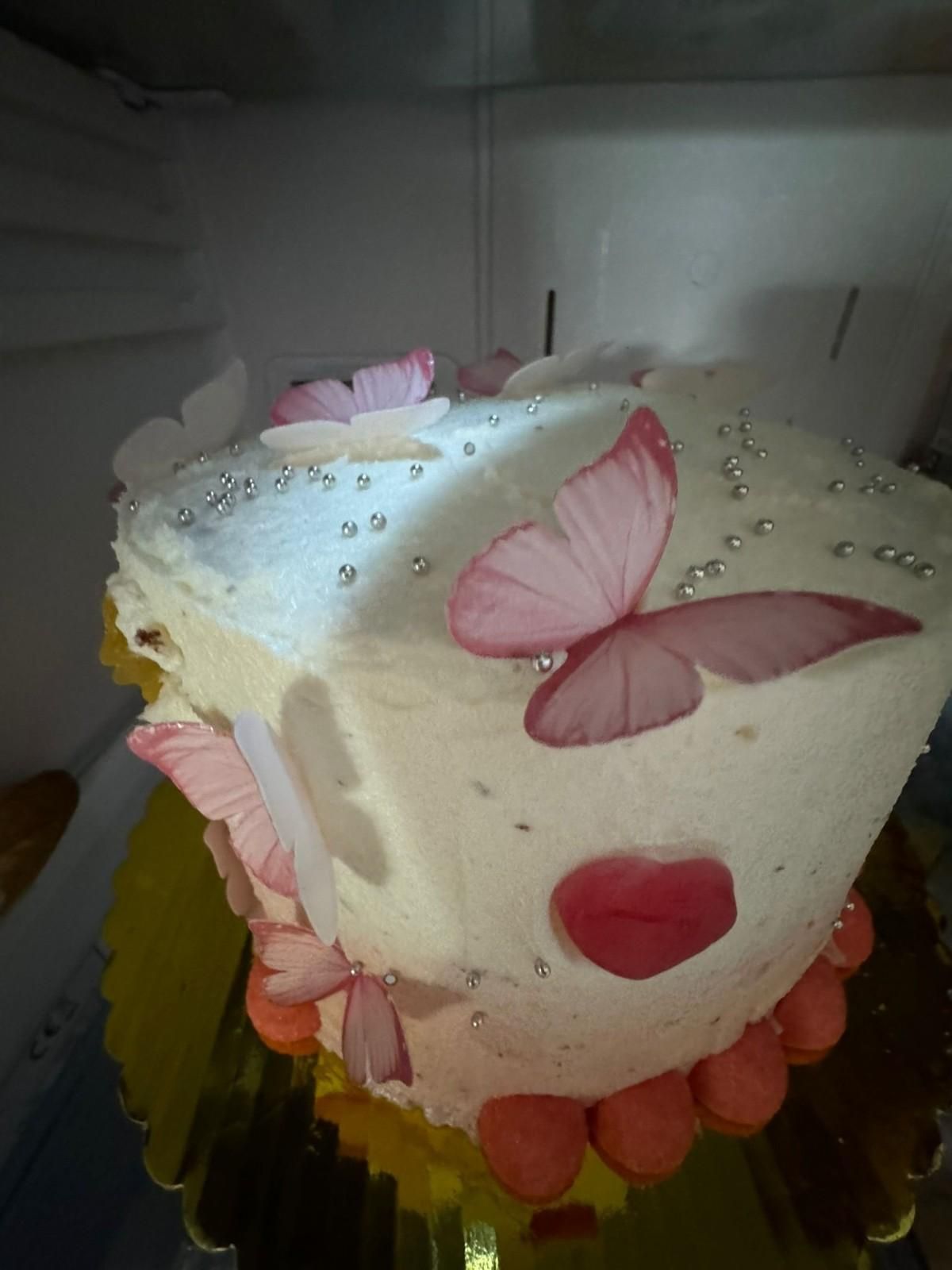 Un pastel blanco glaseado en un refrigerador, decorado con recortes de mariposas rosas, chispas plateadas y detalles de corazones rojos.