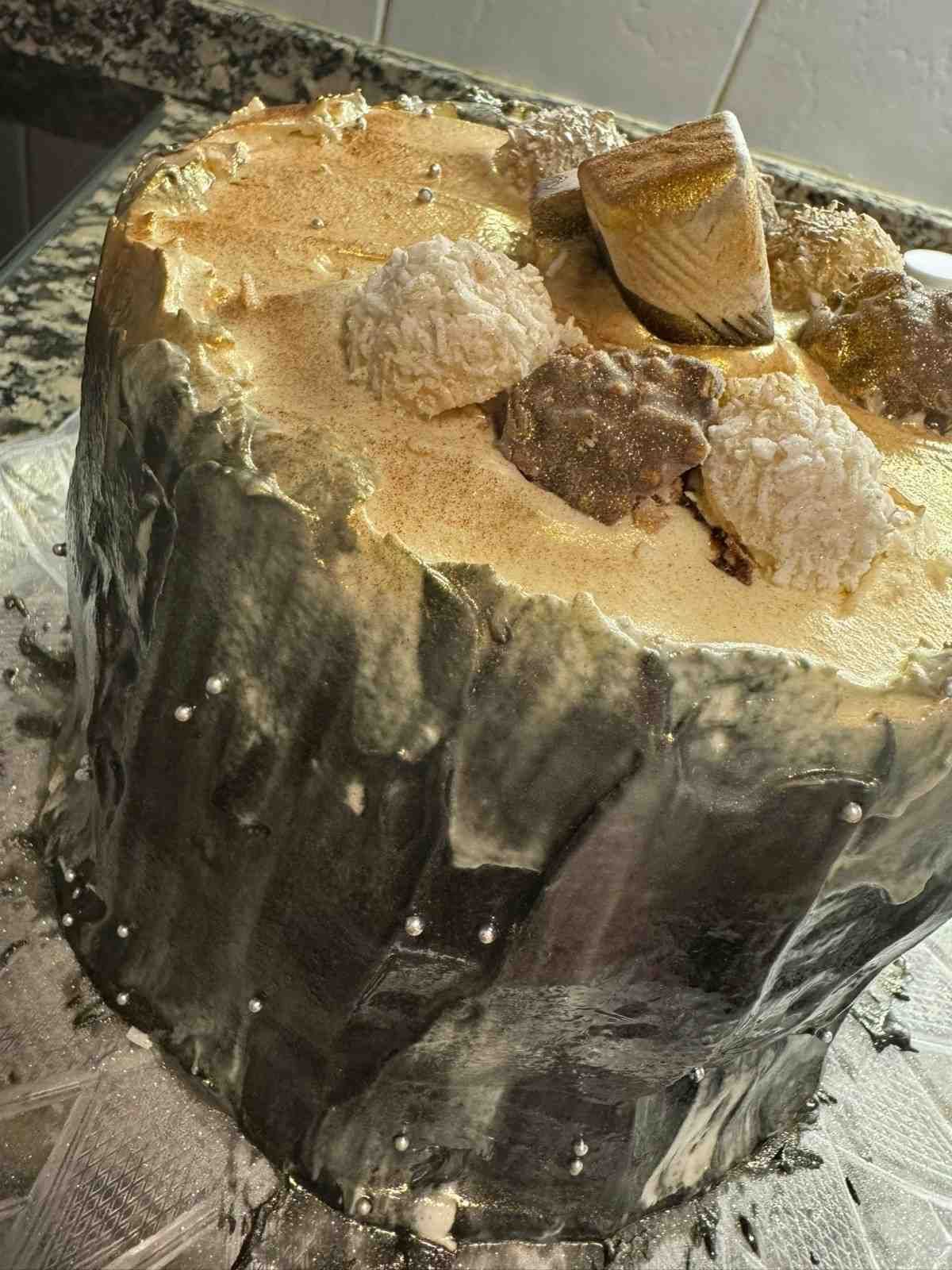 Un pastel redondo con un lado oscuro y texturizado que recuerda a la corteza de un árbol y una parte superior de color marrón claro, decorado con esferas cubiertas de chocolate esparcidas.