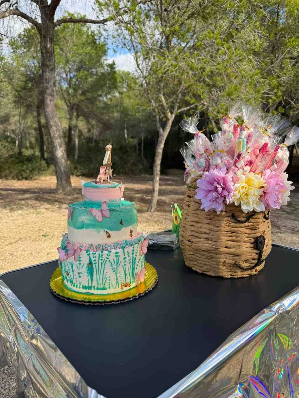 Una tarta de varios pisos decorada con mariposas y una figura de la Torre Eiffel en la parte superior, junto a una cesta con bolsitas de dulces con motivos florales, al aire libre.