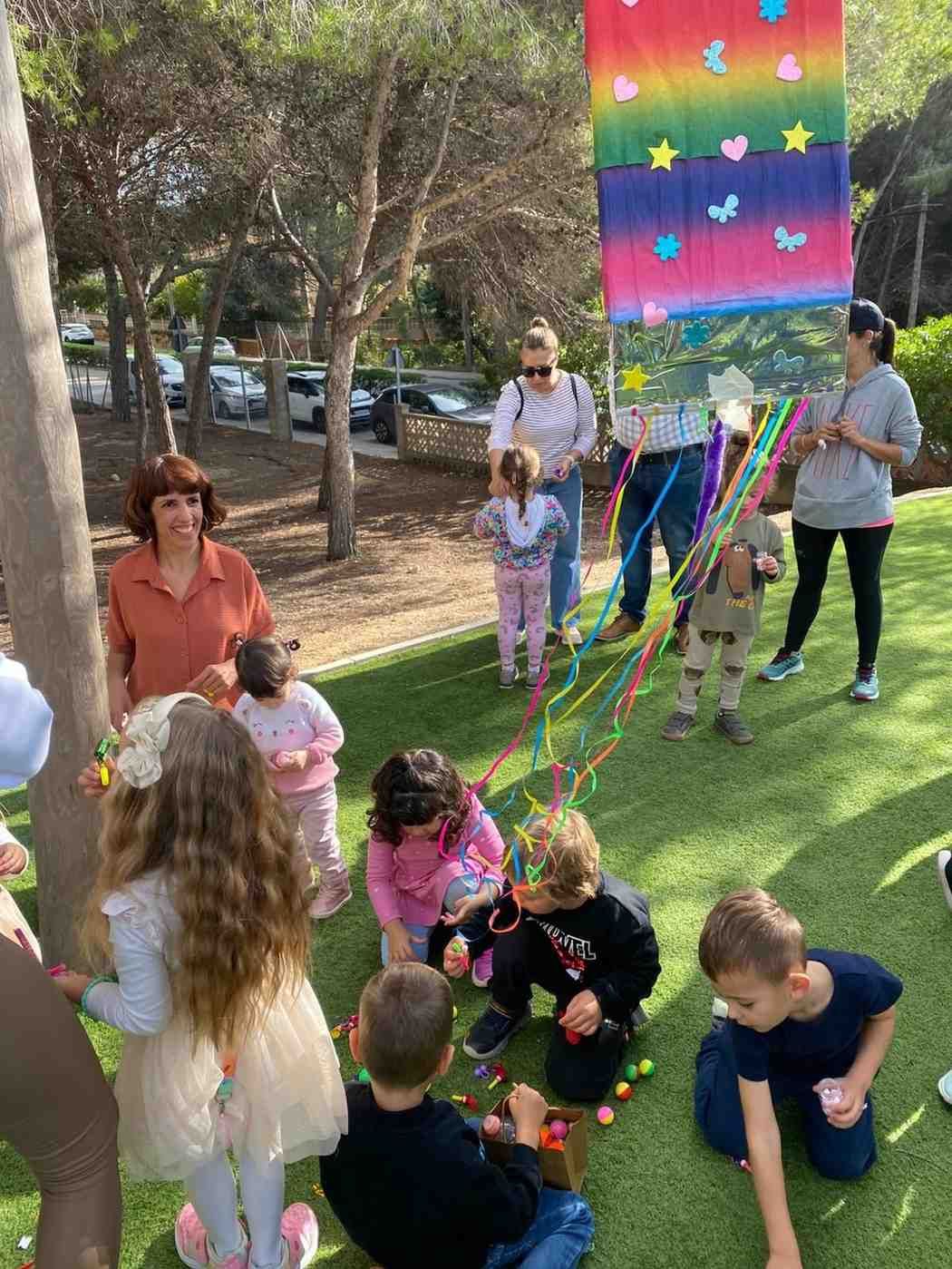 Un grupo de niños y adultos juegan con serpentinas bajo una pancarta con los colores del arcoíris en una zona verde al aire libre.