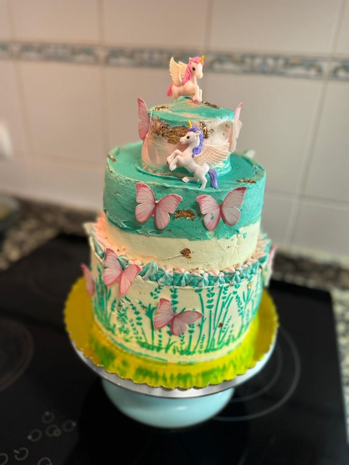 Una tarta de dos pisos decorada con mariposas, un estampado floral con plantilla, detalles dorados y dos adornos de unicornios para la parte superior.