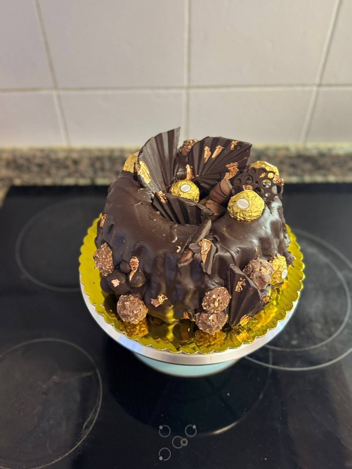 Un pastel de chocolate tipo bundt decorado con bombones Ferrero Rocher y virutas de chocolate sobre una base dorada para pasteles.