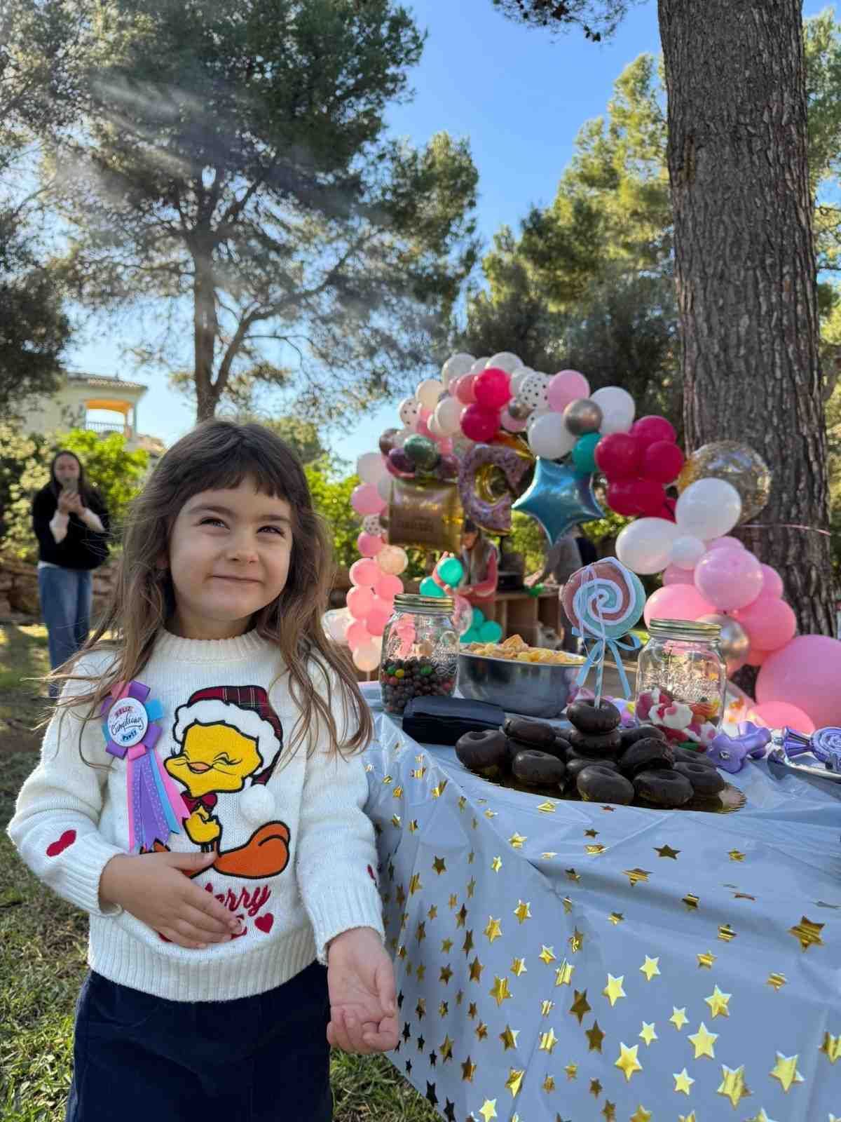 Un niño se encuentra de pie frente a una mesa decorada con un arco de globos y dulces en una fiesta al aire libre.