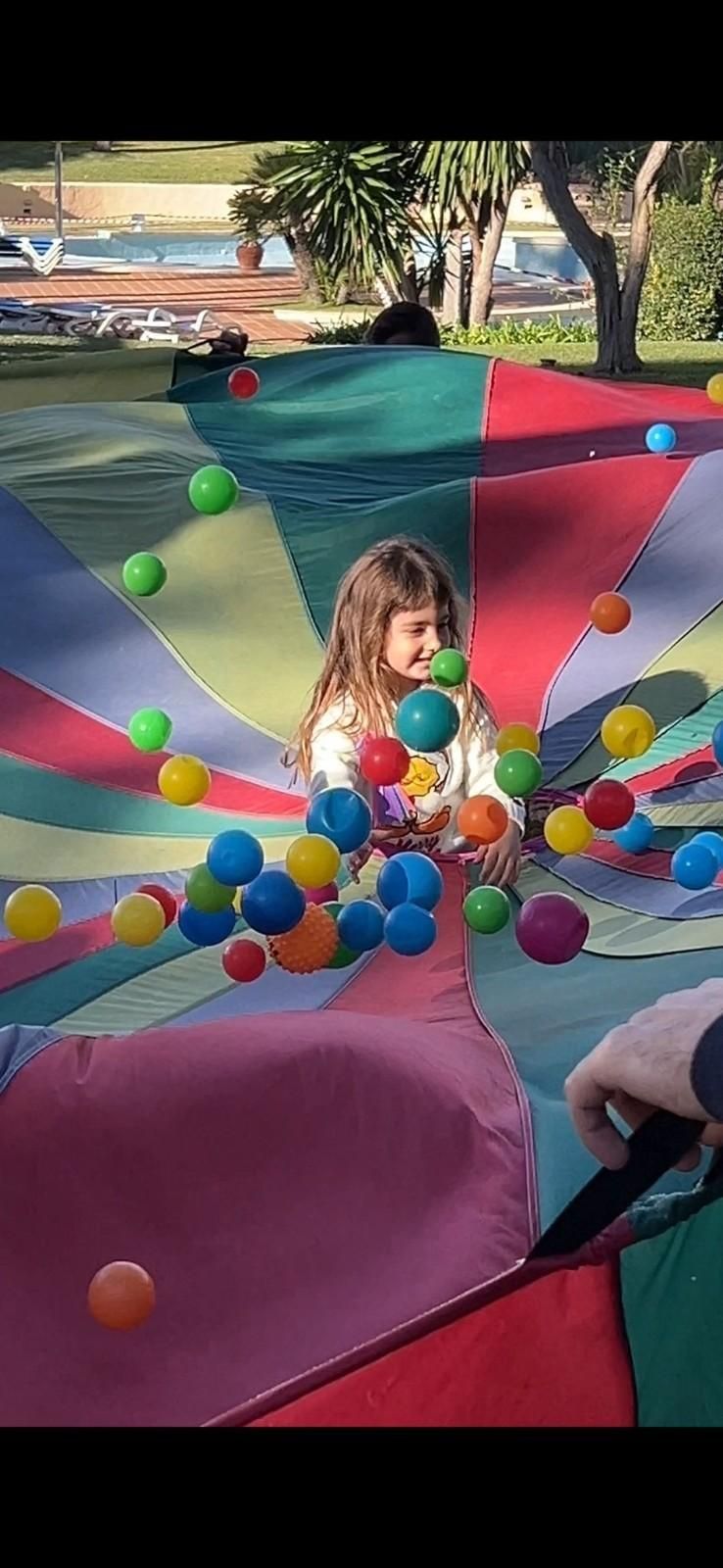 Un niño sonríe mientras juega con coloridas pelotas de plástico en un gran paracaídas multicolor al aire libre.