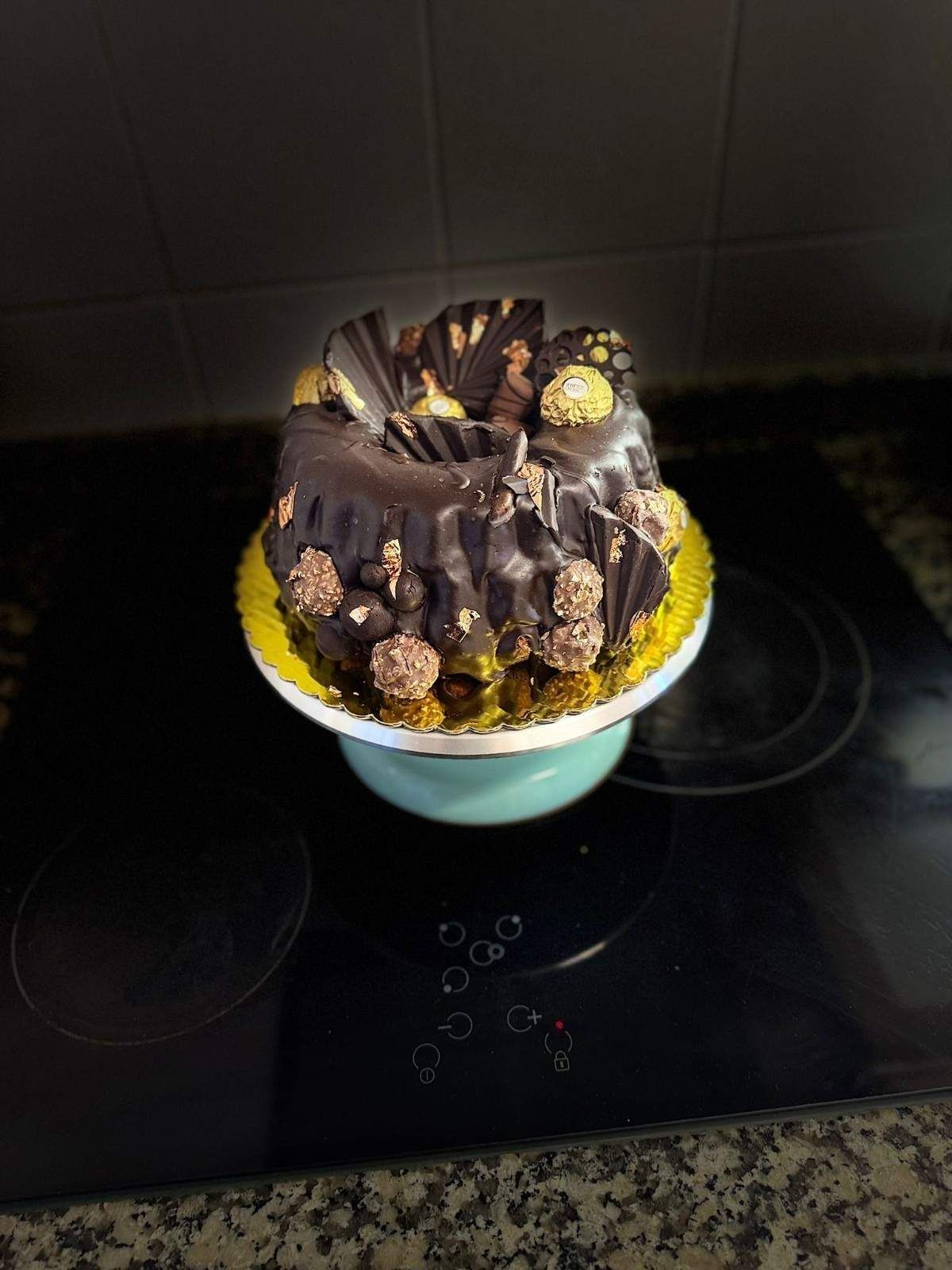 Un bizcocho de chocolate negro tipo bundt, adornado con bombones Ferrero Rocher, sobre una base dorada colocada sobre un soporte para pasteles de color azul claro.