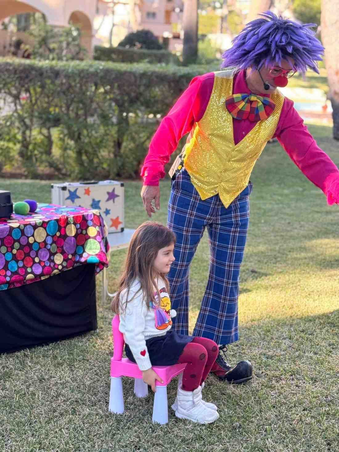 Un payaso con el pelo morado y un chaleco amarillo de lentejuelas actúa para un niño sentado en una silla rosa sobre un césped.