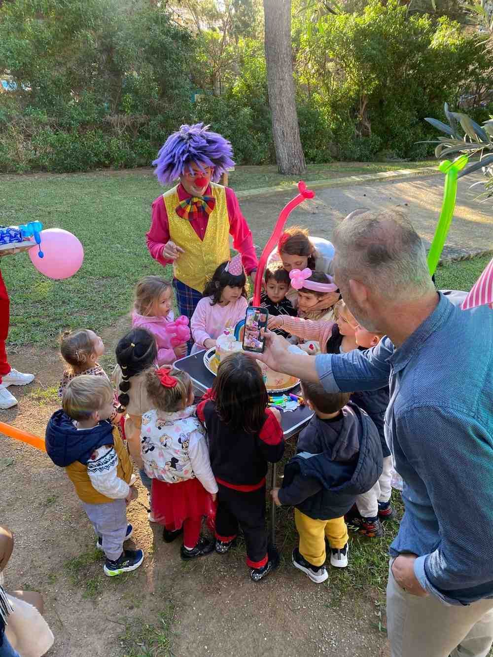 Un payaso con el pelo morado y un chaleco amarillo entretiene a un grupo de niños en una fiesta de cumpleaños al aire libre.