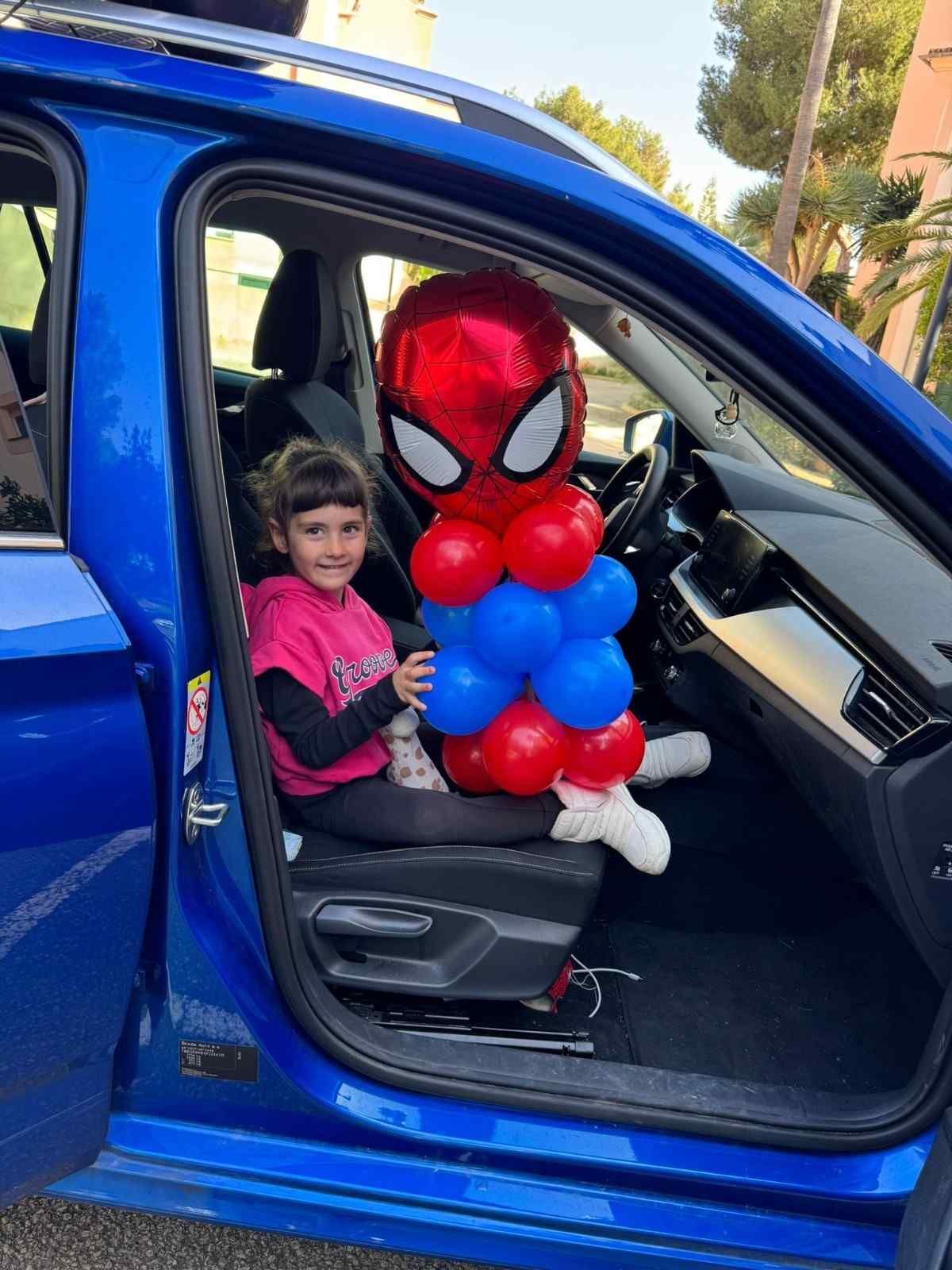 Un niño sonriente está sentado en el asiento del copiloto de un coche azul, junto a un gran globo decorado con la figura de Spiderman.