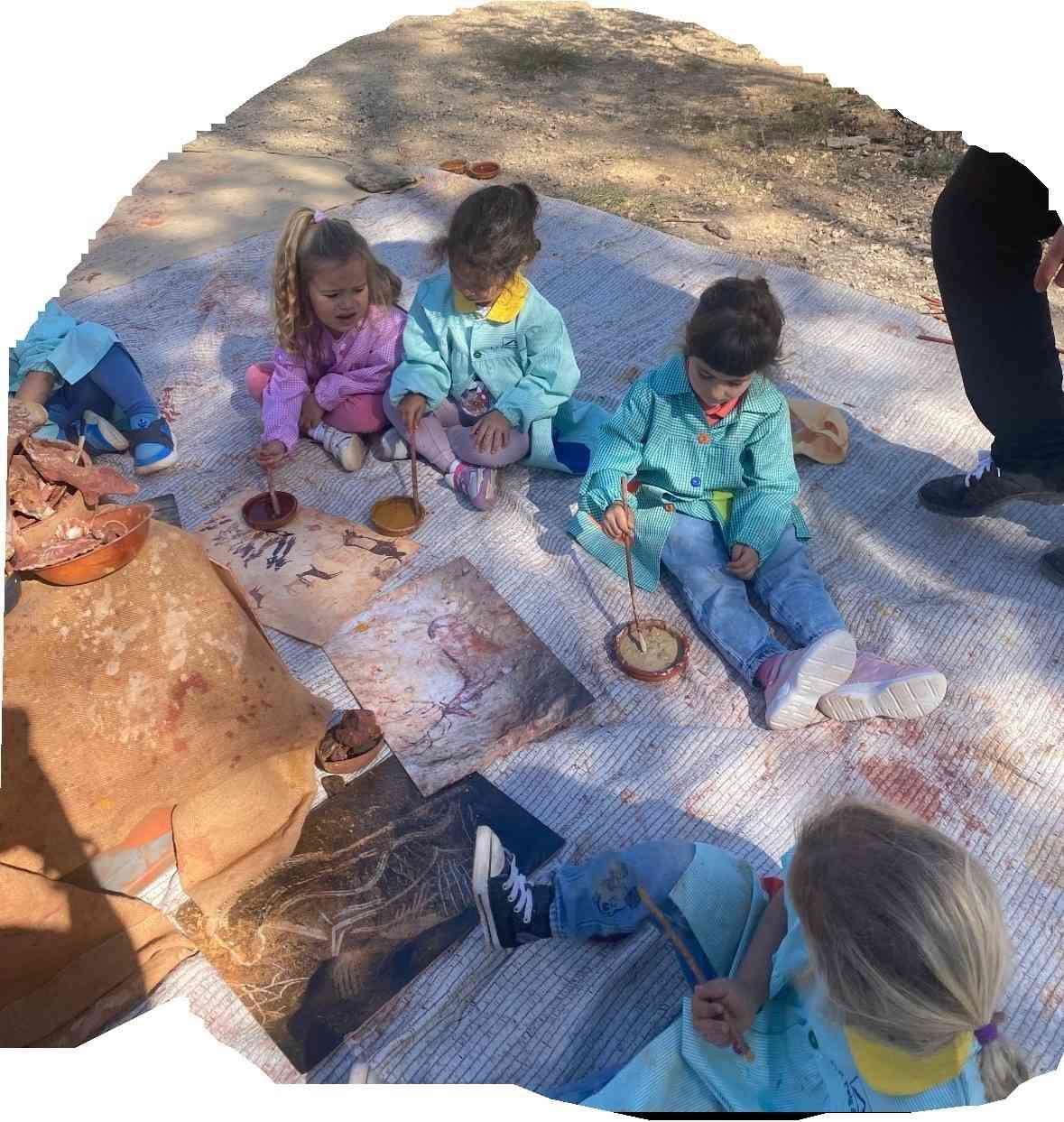 Cuatro niños con delantales están sentados en una esterilla al aire libre, utilizando pintura y arcilla para crear proyectos artísticos sobre papel y discos de arcilla.