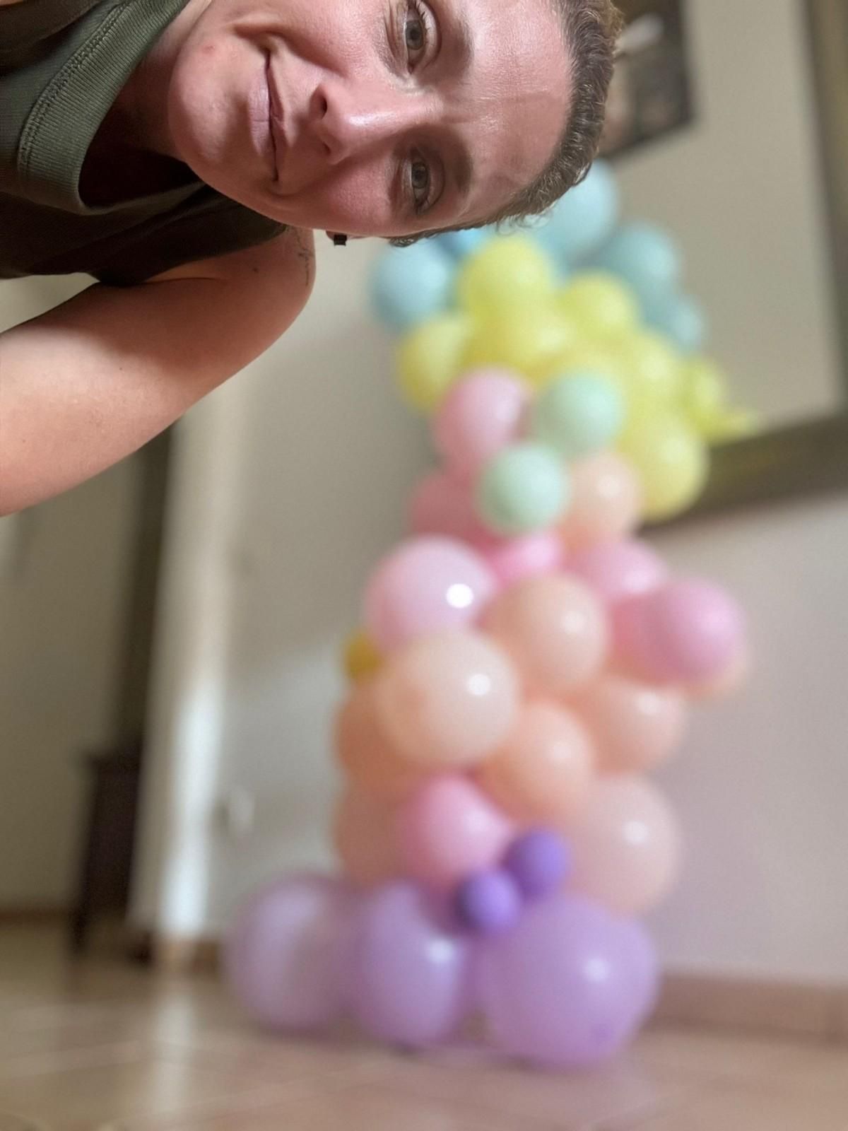 Una persona posando cerca de una torre de globos de colores pastel dentro de una casa.