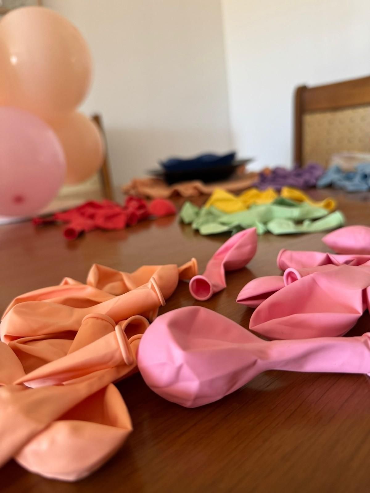 Globos desinflados en tonos rosas, naranjas, verdes y rojos esparcidos sobre una mesa de madera junto a globos inflados.
