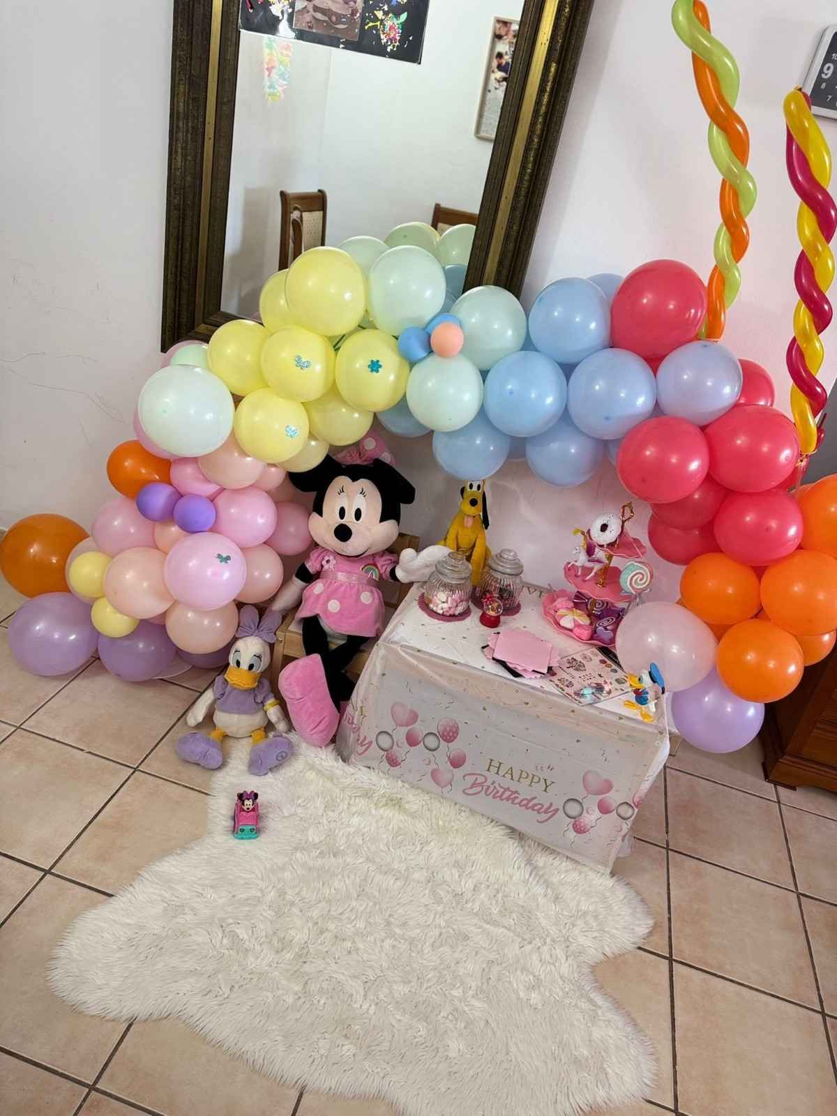 Una decoración de cumpleaños con temática de Minnie Mouse, con globos, un peluche de Minnie y Daisy Duck, y una mesita sobre una alfombra.