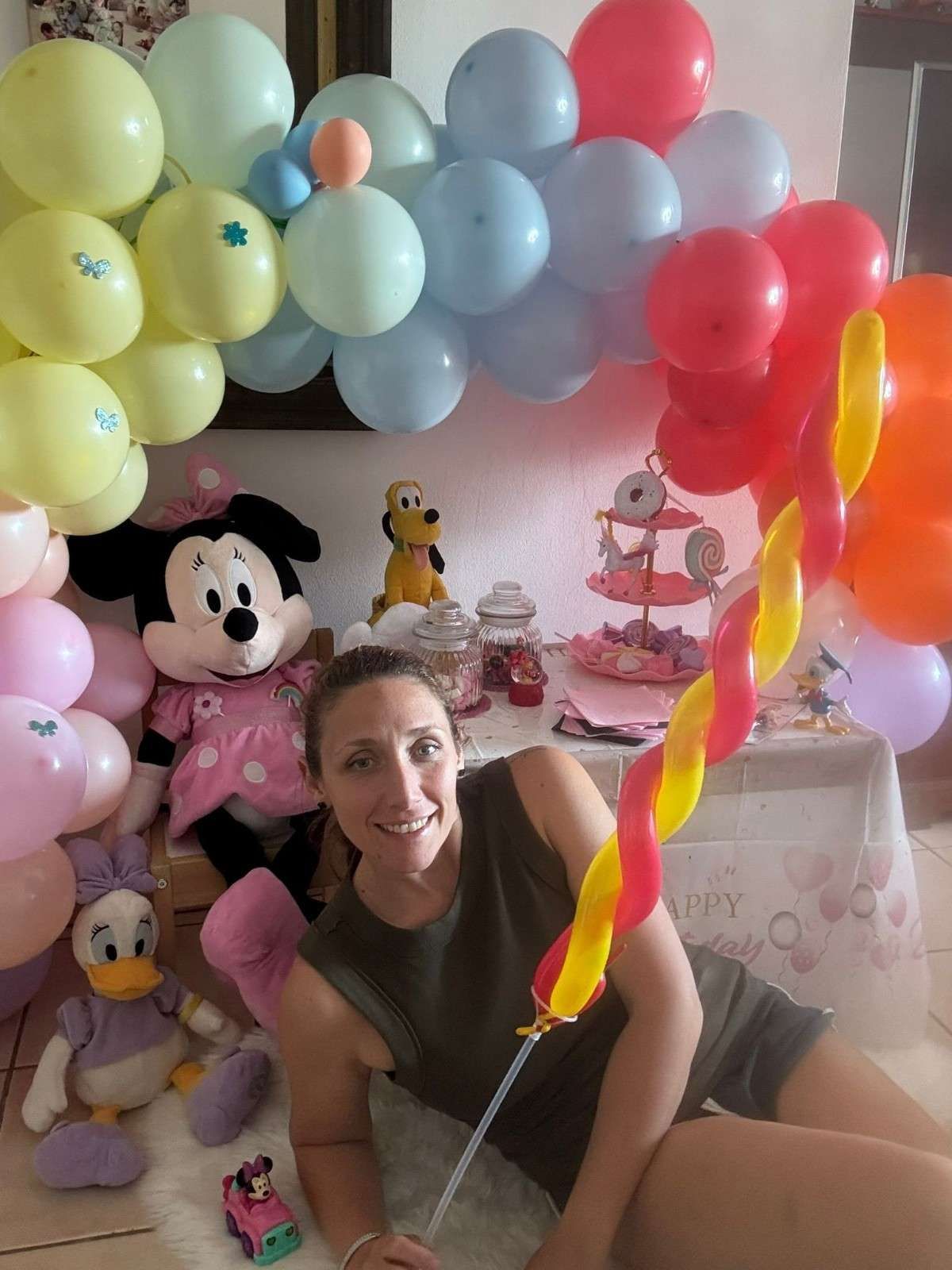 Una persona sonríe frente a una decoración de fiesta de Minnie Mouse que incluye un colorido arco de globos, peluches y una mesa.