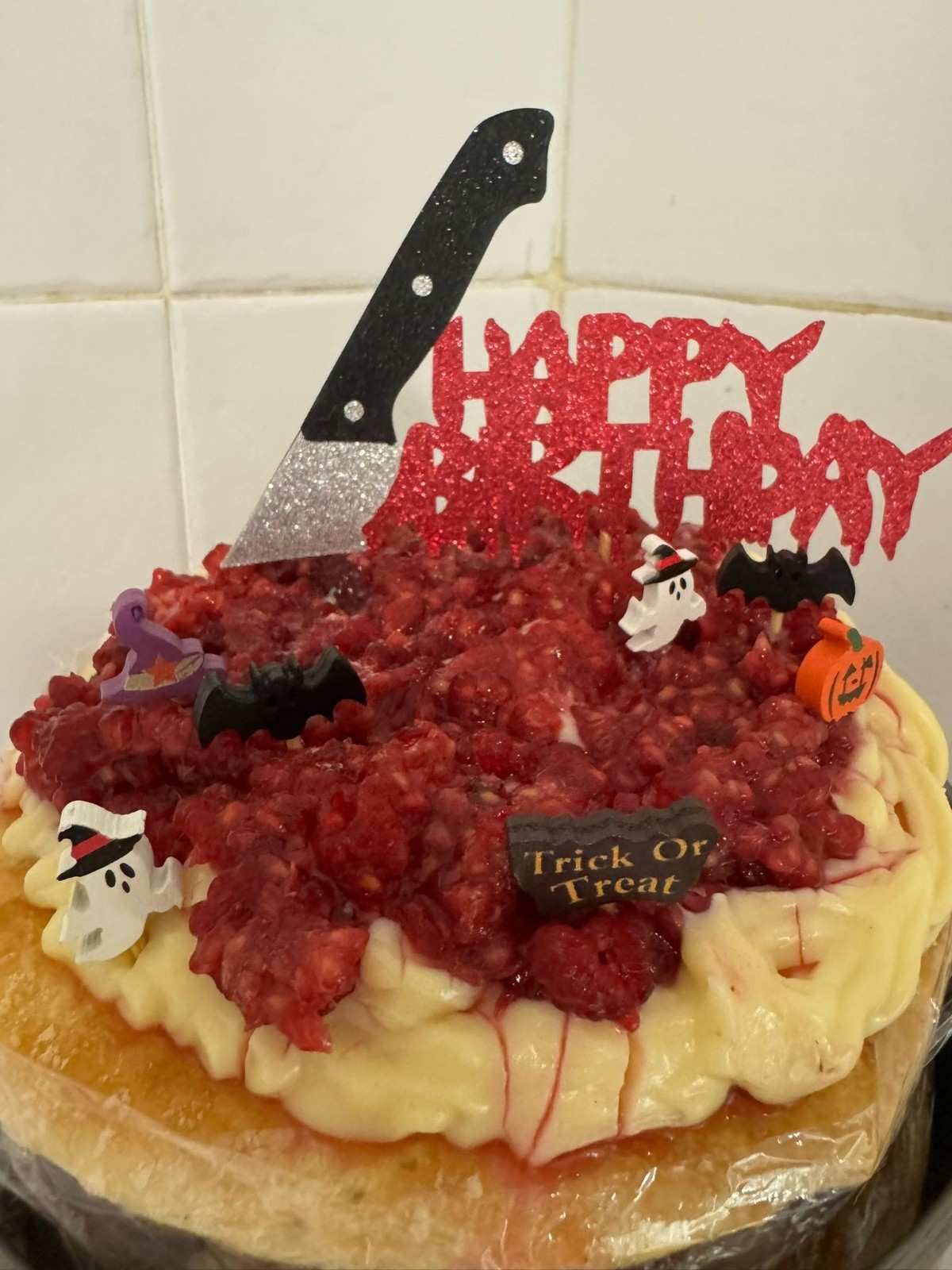 Una tarta de cumpleaños decorada con fruta roja, un adorno en forma de cuchillo y pequeñas decoraciones de plástico con temática de Halloween.