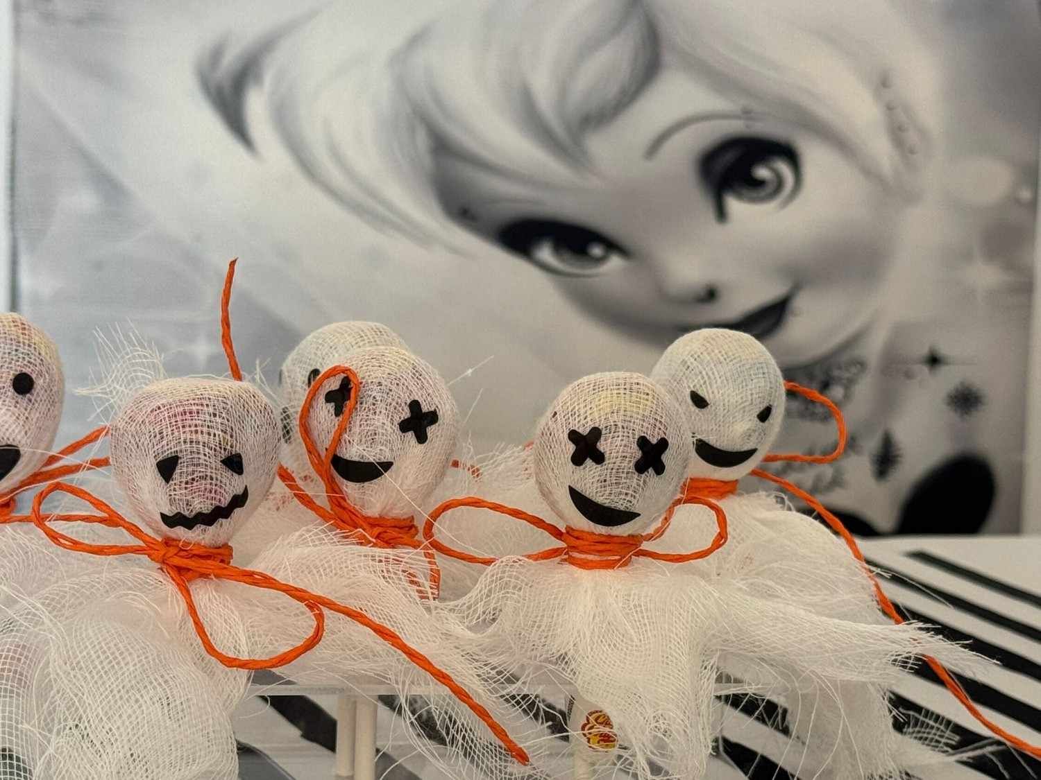 Varios pequeños fantasmas blancos con forma de piruleta, con caras dibujadas y atados con cordel naranja, se encuentran frente a un fondo con la imagen de una cara de dibujos animados.