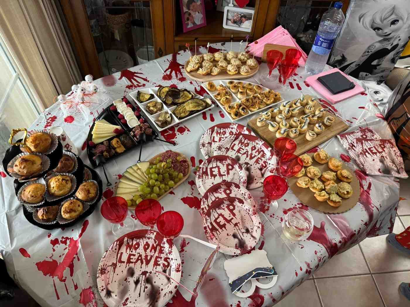 Una mesa de comedor con temática de terror cubierta de salpicaduras de sangre falsa, con bandejas de comida y platos impresos con la frase 