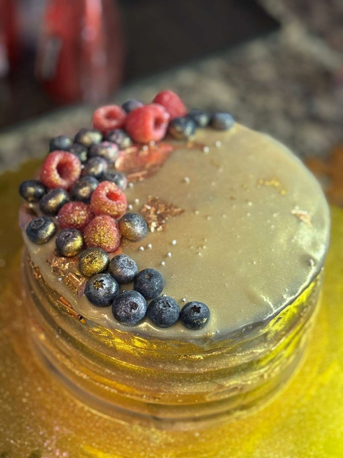 Un pastel de varias capas con un glaseado dorado brillante, decorado con frambuesas frescas, arándanos y detalles de pan de oro.