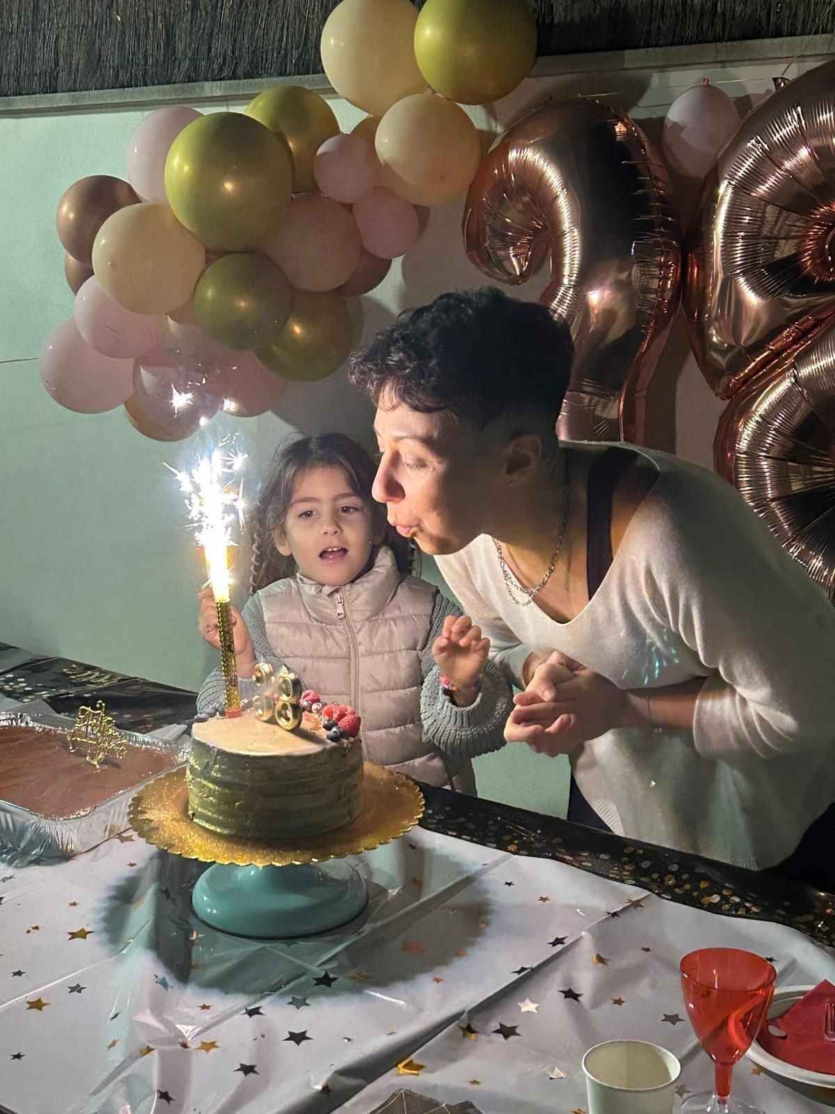 Una persona sopla una vela de bengala en una tarta de cumpleaños junto a un niño, rodeados de globos dorados y rosas.