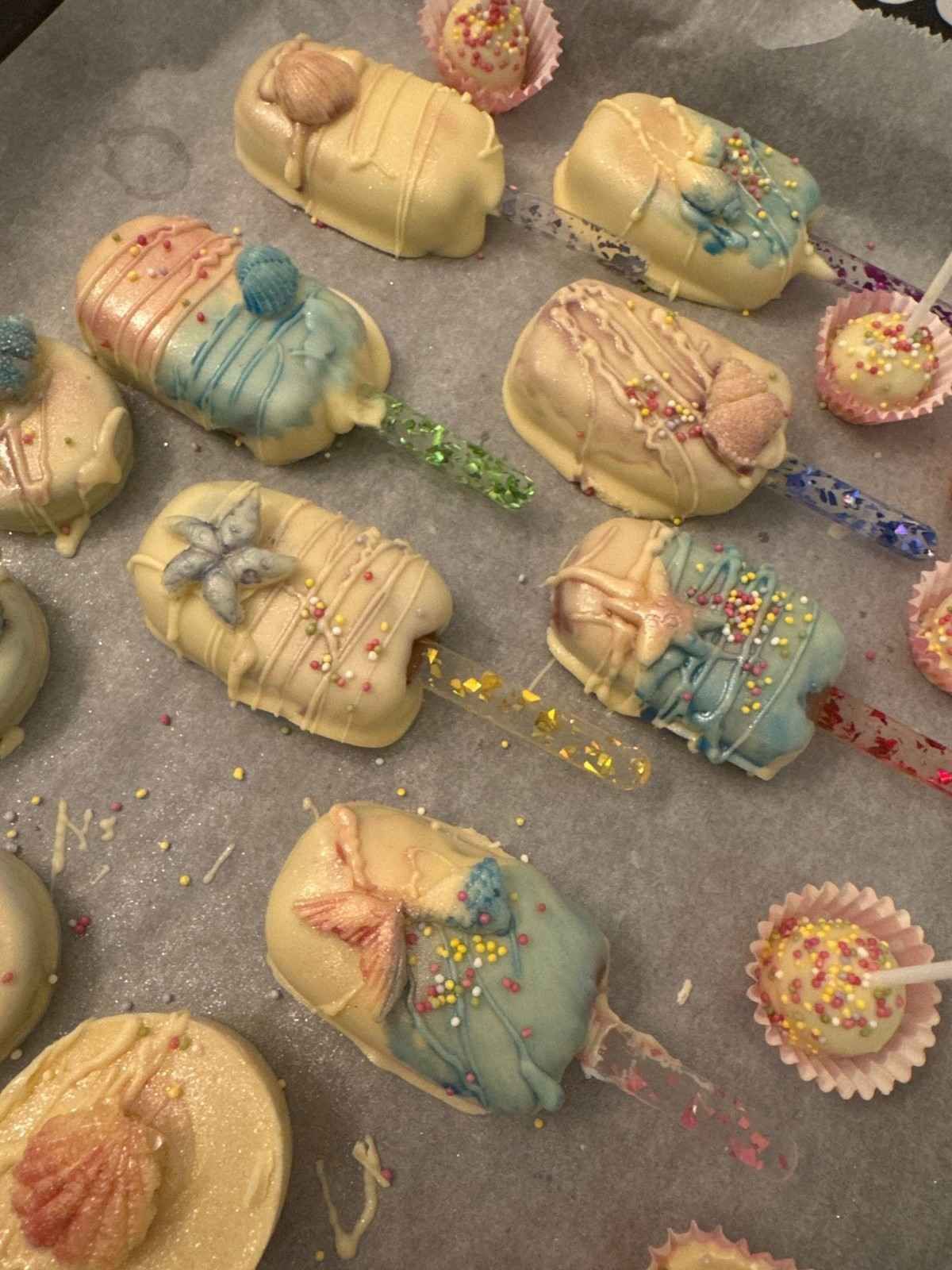 Cake pops caseros con forma de helado, decorados con detalles en azul pastel, blanco y dorado sobre papel pergamino.