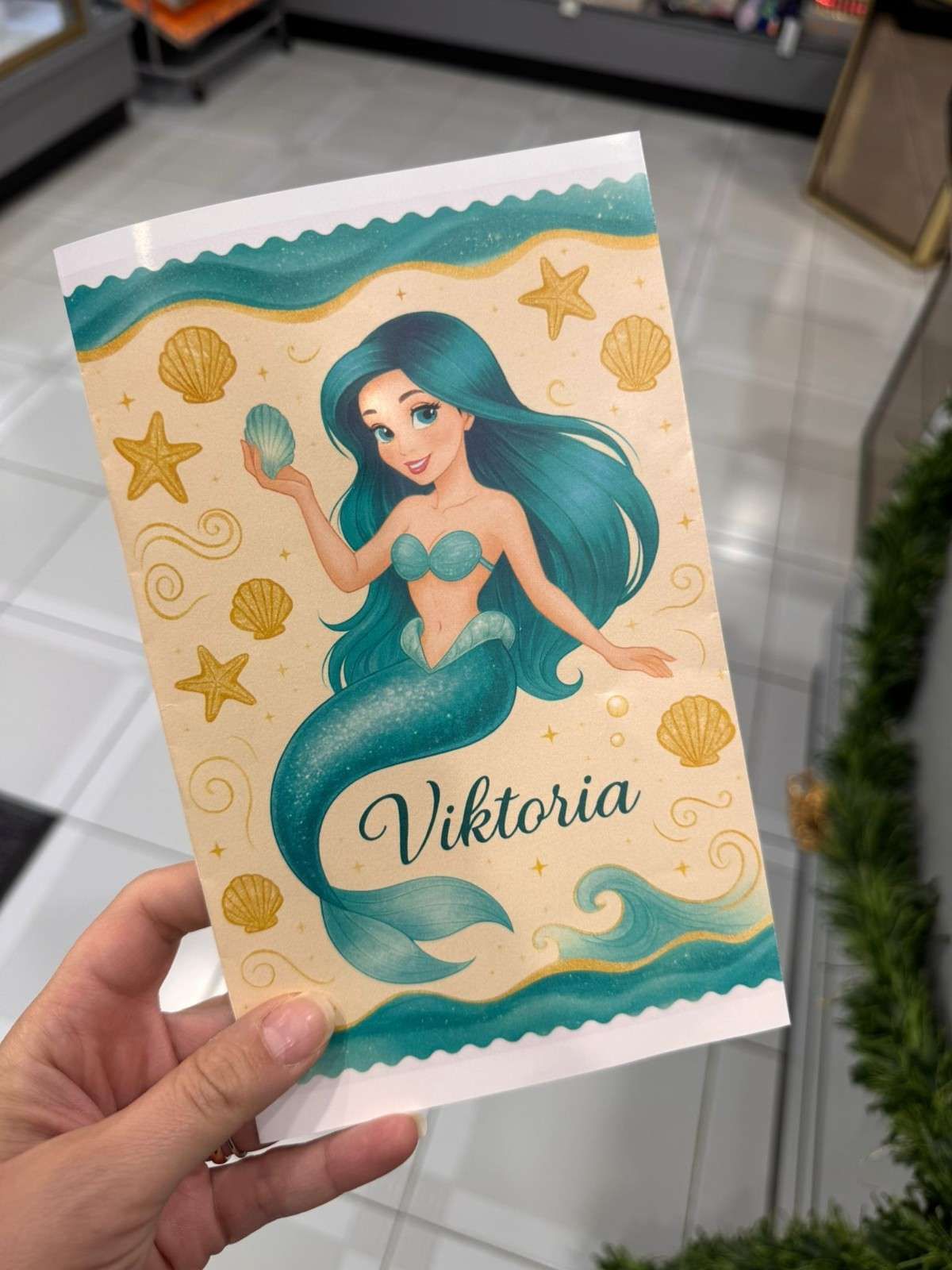 Una mano sostiene una tarjeta personalizada con la ilustración de una sirena de cabello azul ondulado y el nombre 
