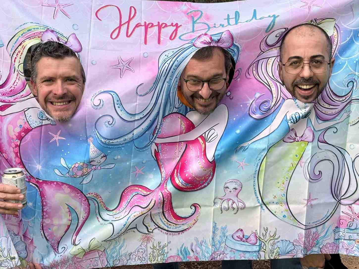 Tres personas sonríen a través de recortes de rostros en una colorida pancarta con temática de sirenas que dice 