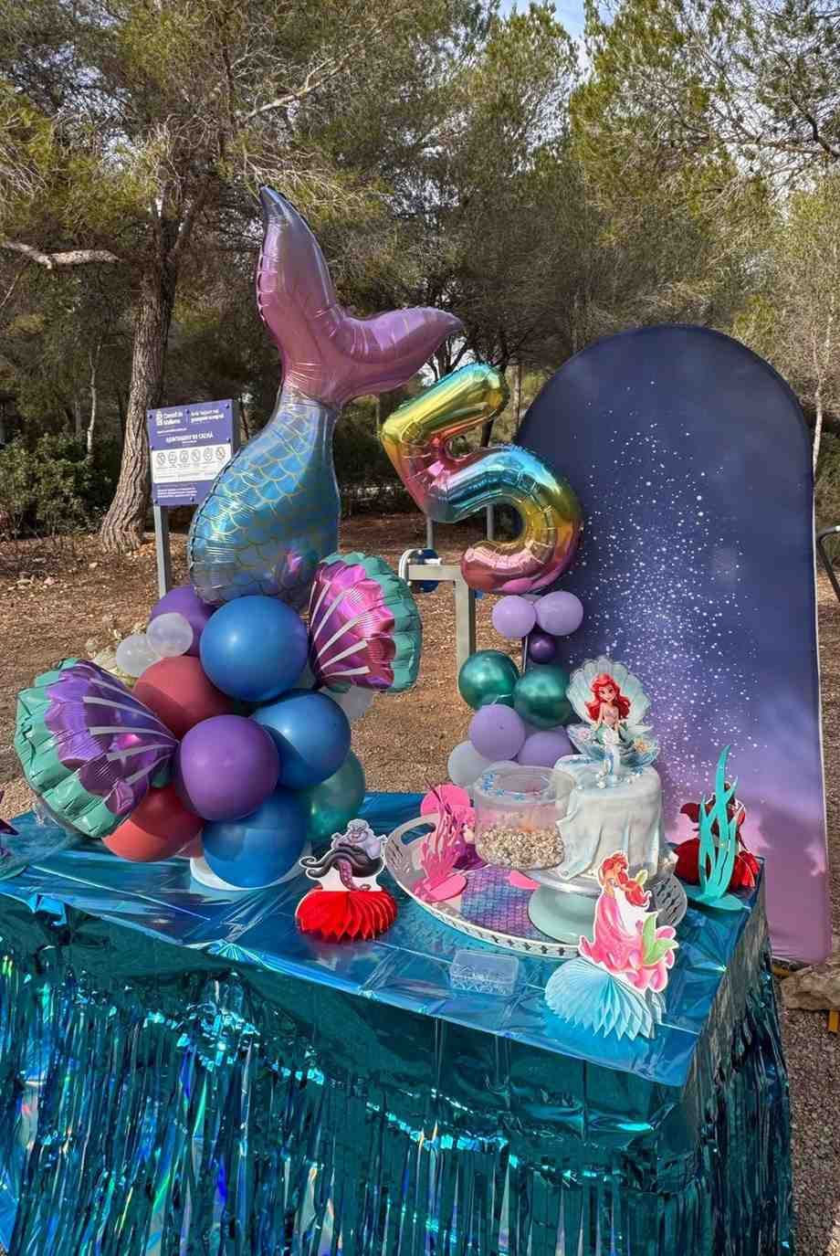 Una decoración de fiesta con temática de sirenas sobre una mesa con flecos azules, con un globo en forma de cola de sirena, un globo con el número cinco y un pastel.