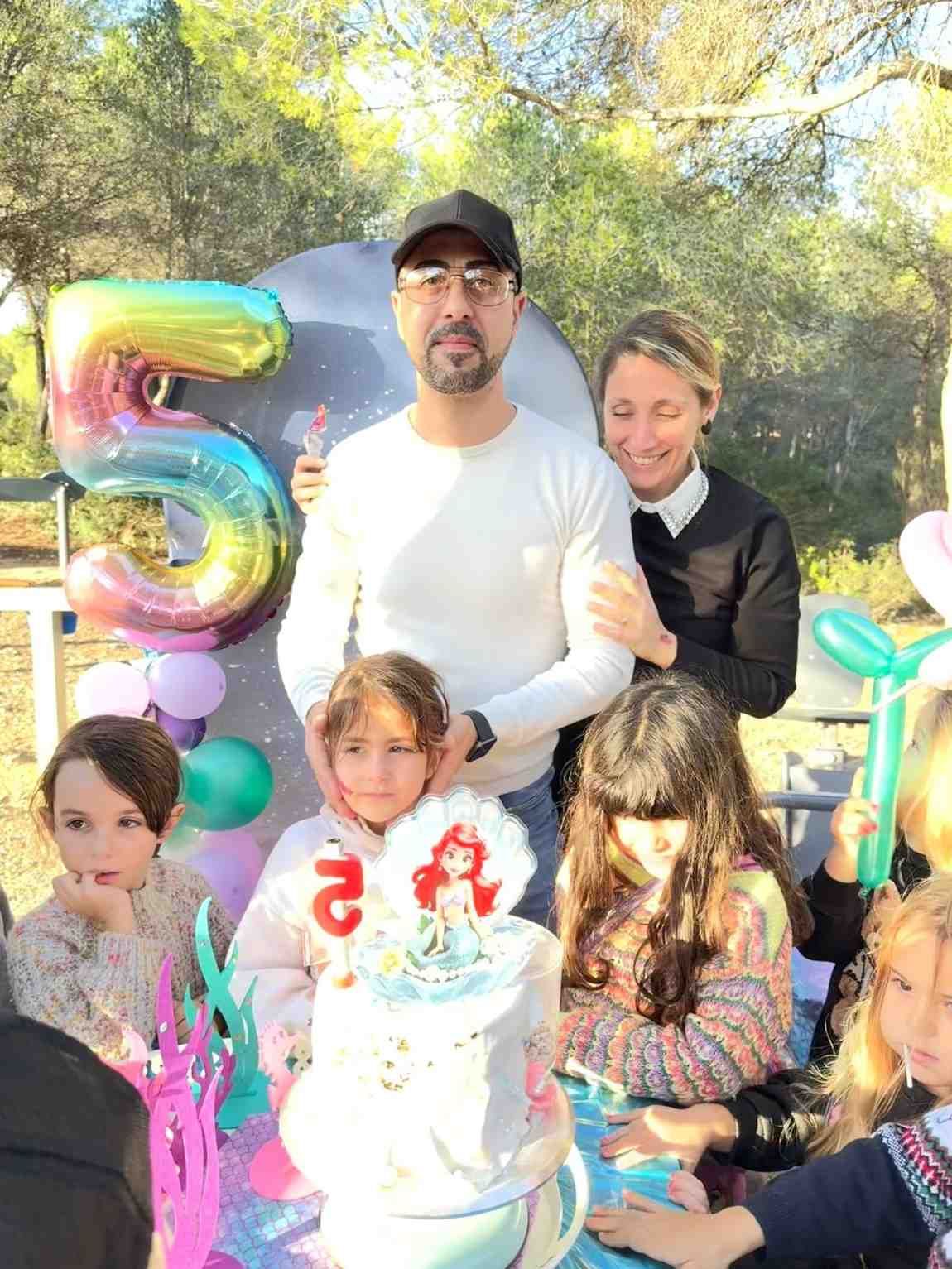 Una familia celebra el quinto cumpleaños de su hijo con pastel, globos y decoraciones al aire libre.