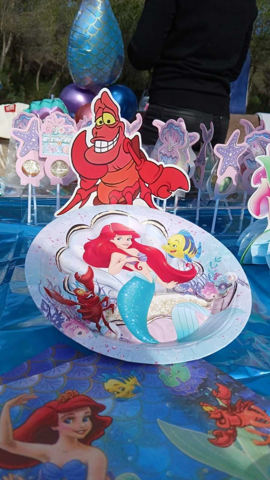 Una mesa decorada con temática de La Sirenita, con platos y adornos de Ariel y Sebastián sobre un mantel azul.