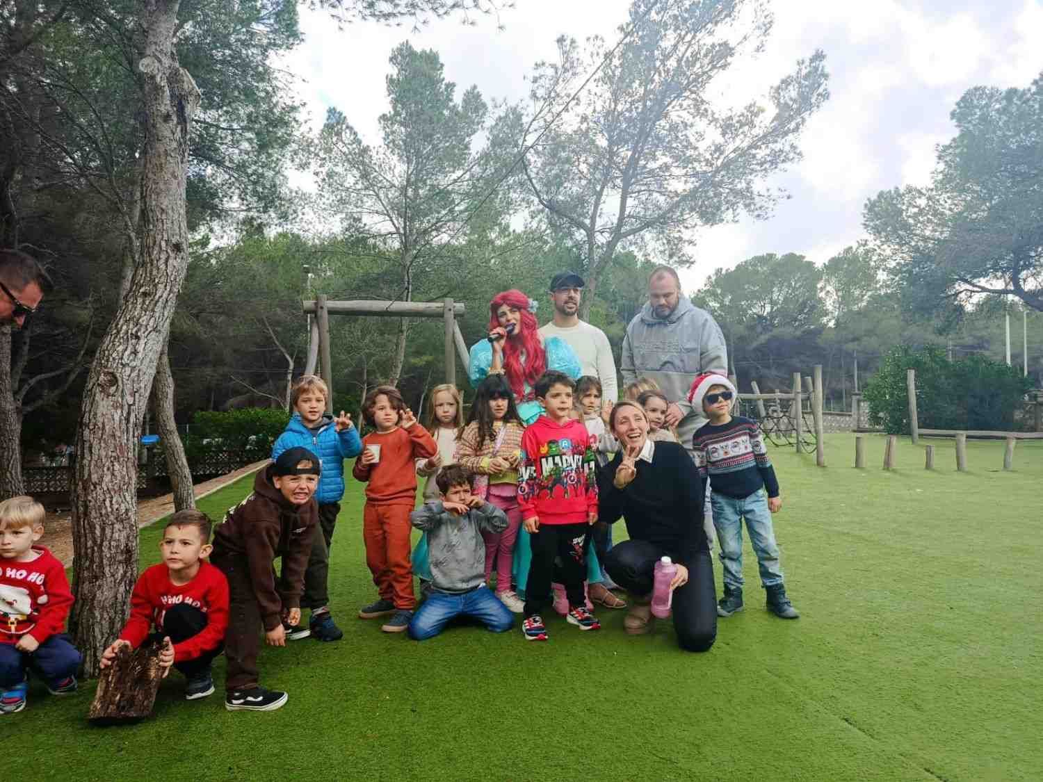 Un grupo de niños y adultos posan juntos al aire libre sobre un césped durante un evento en el que participa un personaje disfrazado.