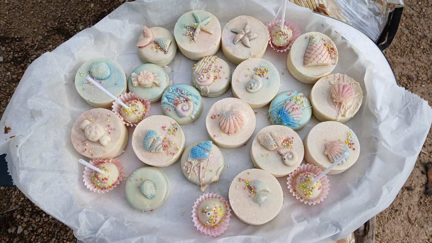 Una bandeja de dulces redondos cubiertos de chocolate blanco y decorados con coloridas formas de conchas marinas y estrellas de mar con temática oceánica.