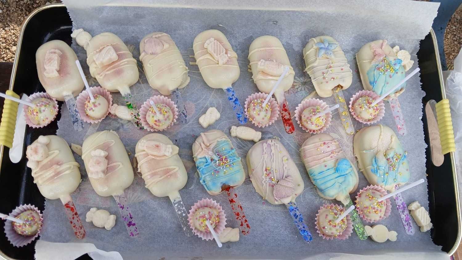 Una bandeja repleta de paletas de pastel decoradas en blanco, azul y rosa, acompañadas de pequeños dulces redondos en vasitos de papel.