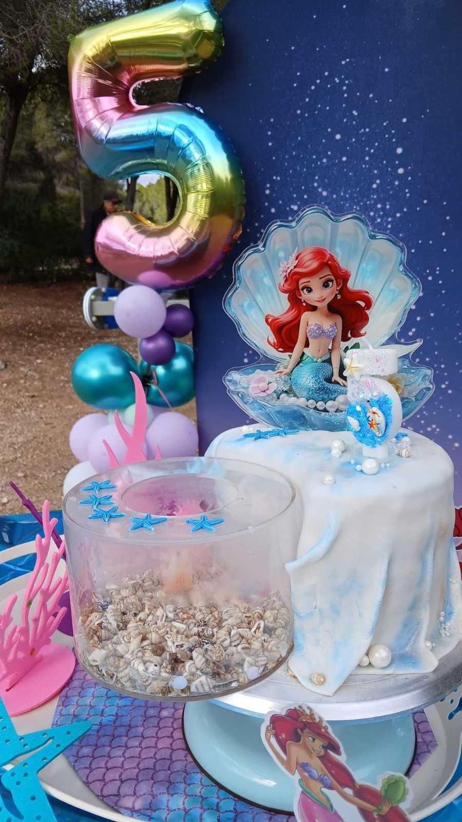 Una decoración de cumpleaños con temática de La Sirenita, con un pastel con la figura de Ariel, una concha marina, un recipiente con motivos oceánicos y el número 5.