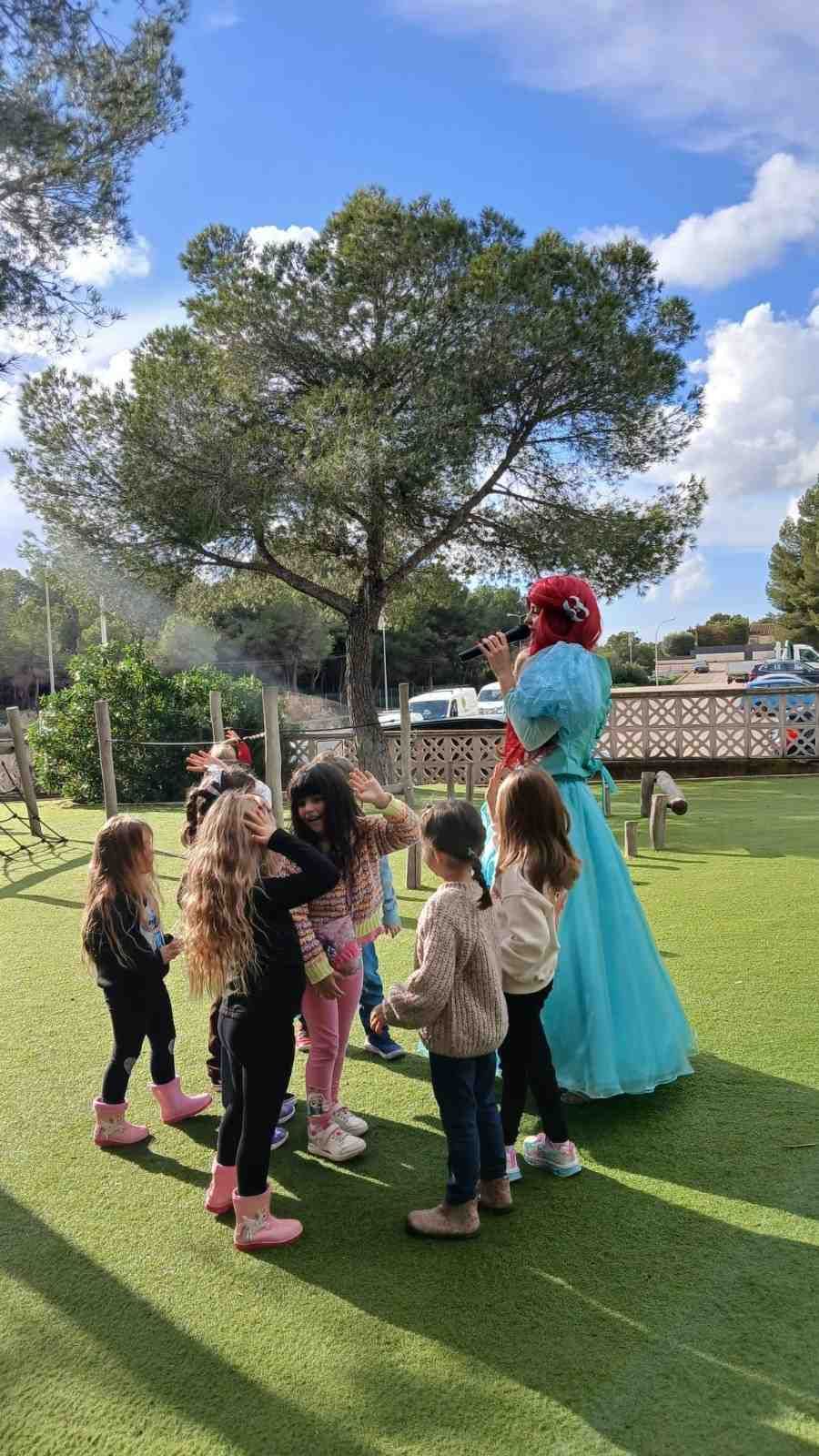 Una animadora disfrazada con un vestido azul interactúa con un grupo de niños en un soleado parque infantil al aire libre, rodeado de vegetación.