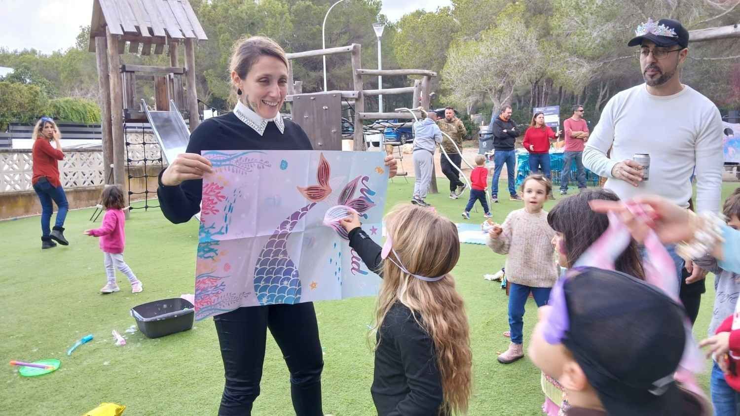Un adulto sostiene un cartel colorido para que un niño lo toque, mientras otros niños se reúnen en un patio de juegos soleado.