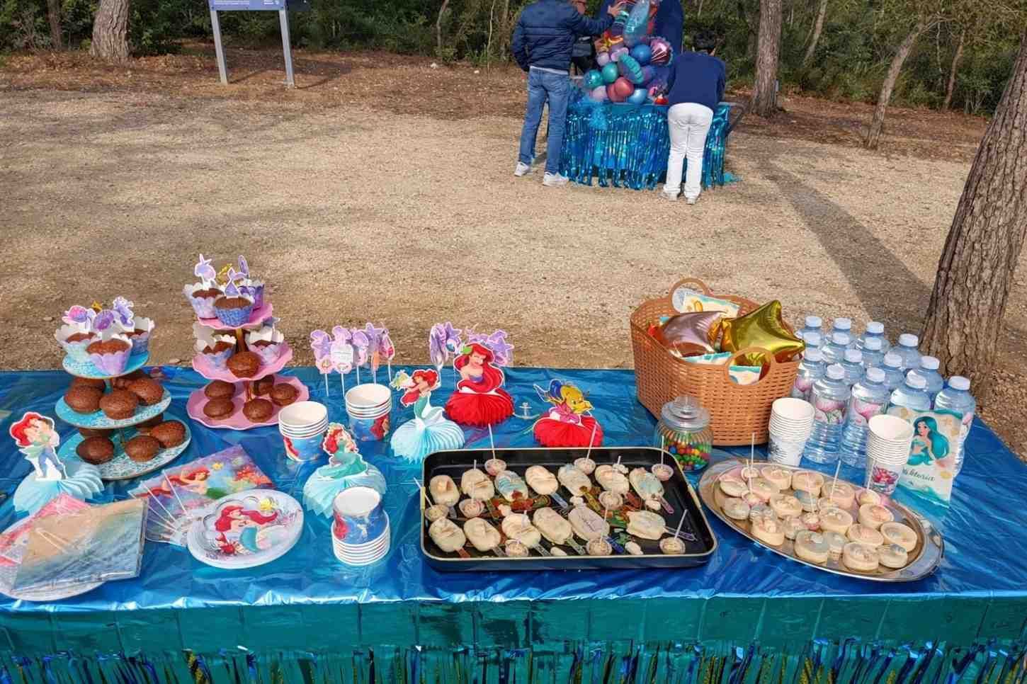 Una mesa de fiesta al aire libre cubierta con un mantel azul, con aperitivos, decoraciones con temática de sirenas y botellas de agua, cerca de una exhibición de globos.