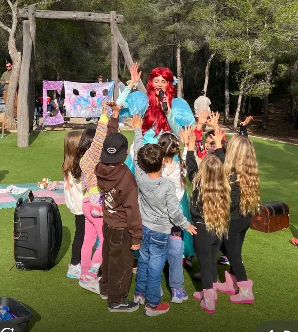 Una persona vestida de sirena con el pelo rojo brillante actúa para un grupo de niños en un parque al aire libre con césped.