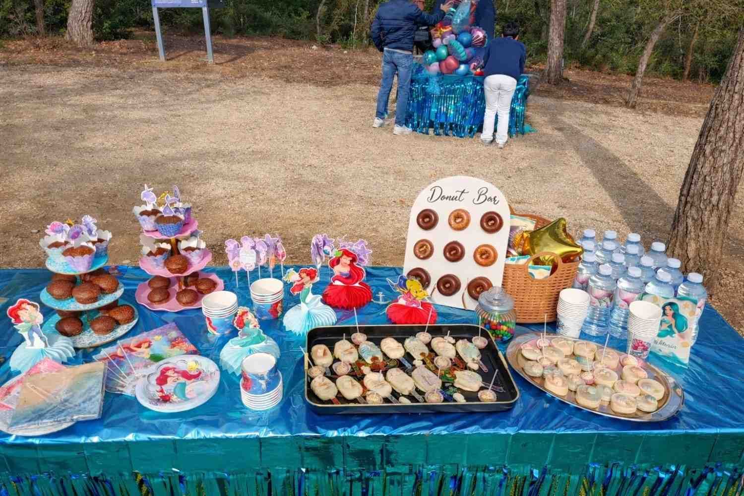 Una mesa de cumpleaños con temática de sirenas en tonos azules, con cupcakes, una bandeja de donas y dulces, al aire libre, con dos personas al fondo.